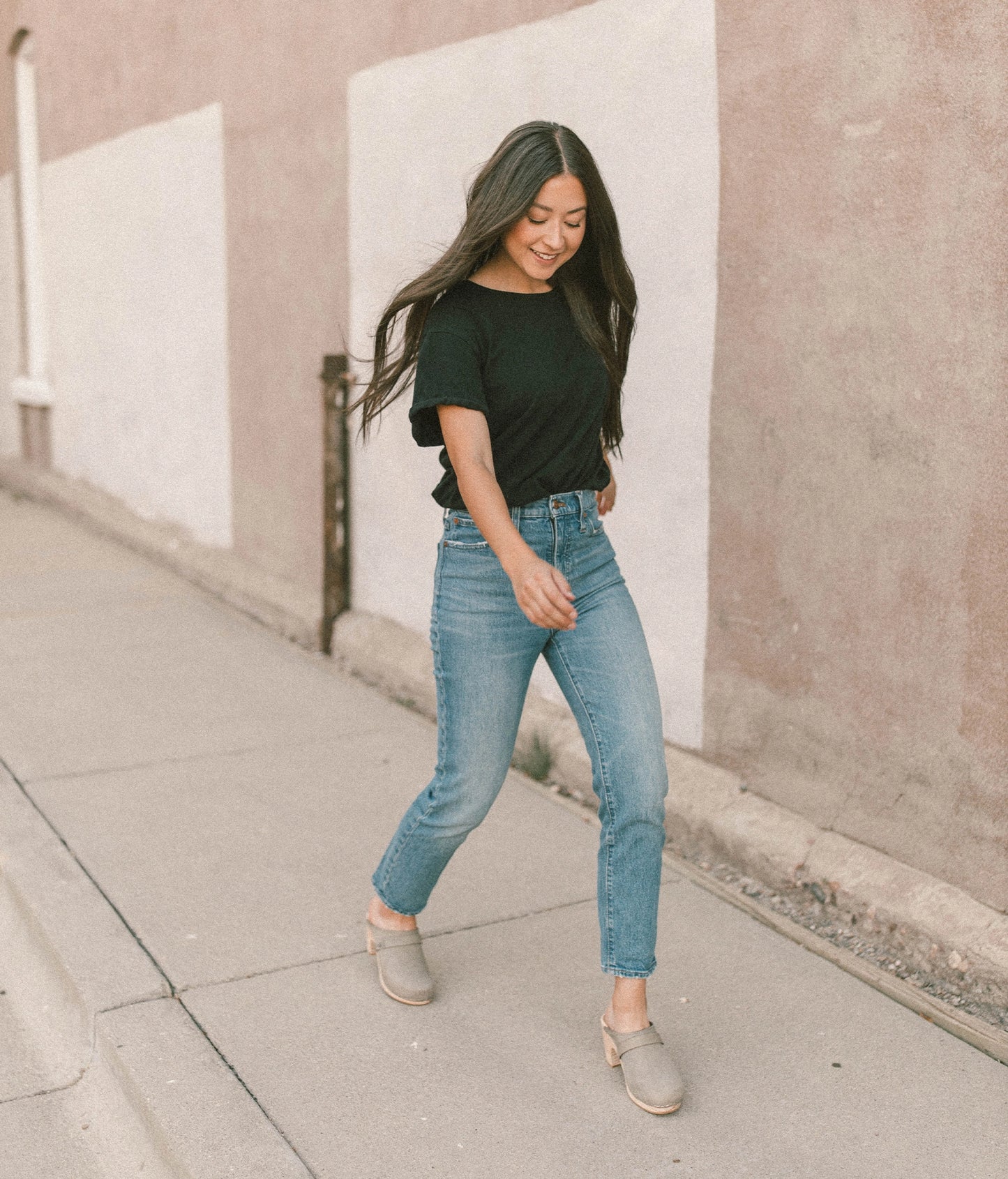 woman walking down the street wearing blue jeans, a black t-shirt with high rise clog mules in stone grey nubuck leather on a light wooden base