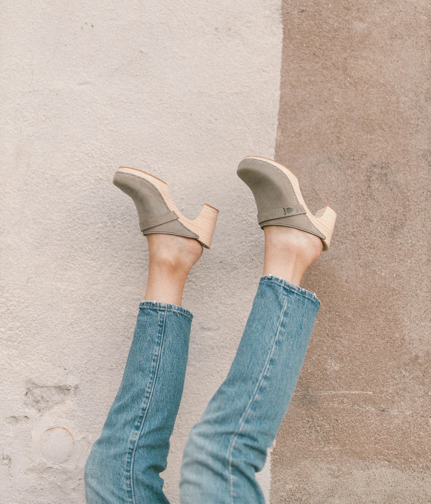 woman holding up her legs in the air while wearing blue jeans with high rise clog mules in stone grey nubuck leather on a light wooden base