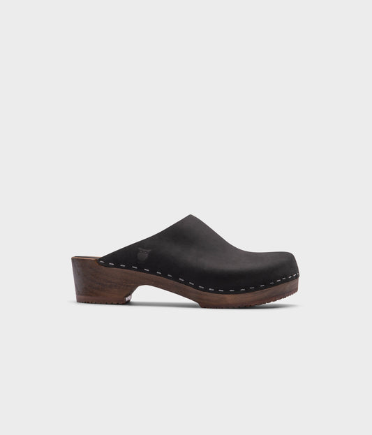 low heeled minimalistic clog mules in black nubuck leather stapled on a dark wooden base