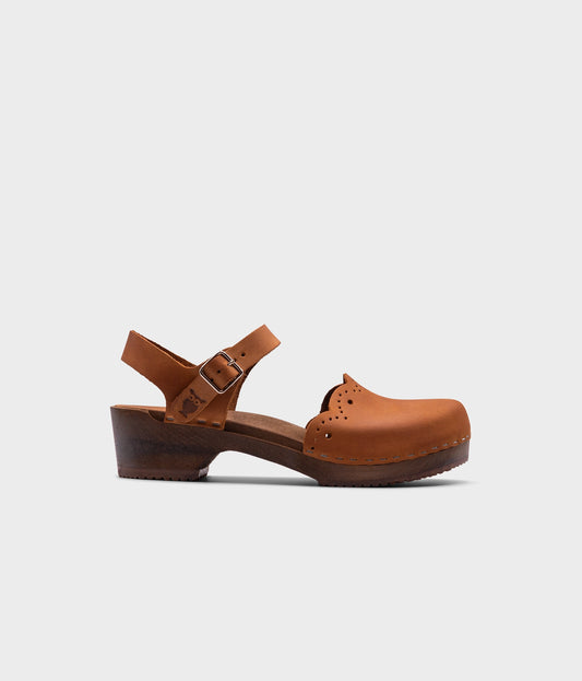 low heeled closed-toe clog sandals in light brown nubuck leather with a wavy leather cutout stapled on a dark wooden base