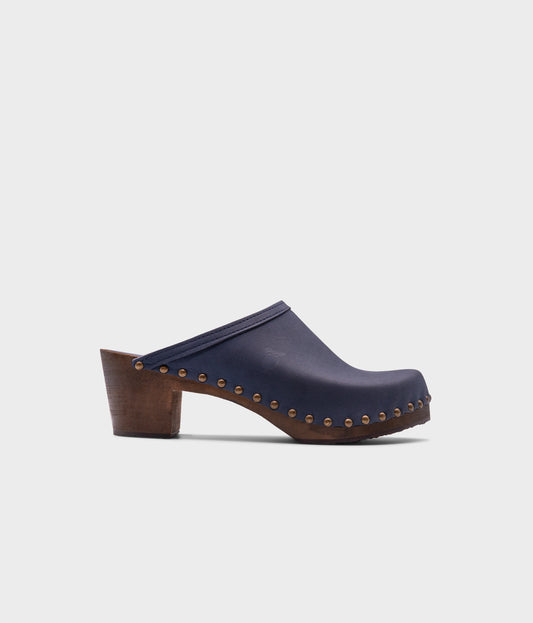 high heeled clog mules in navy blue nubuck leather stapled on a dark wooden base with brass gold studs