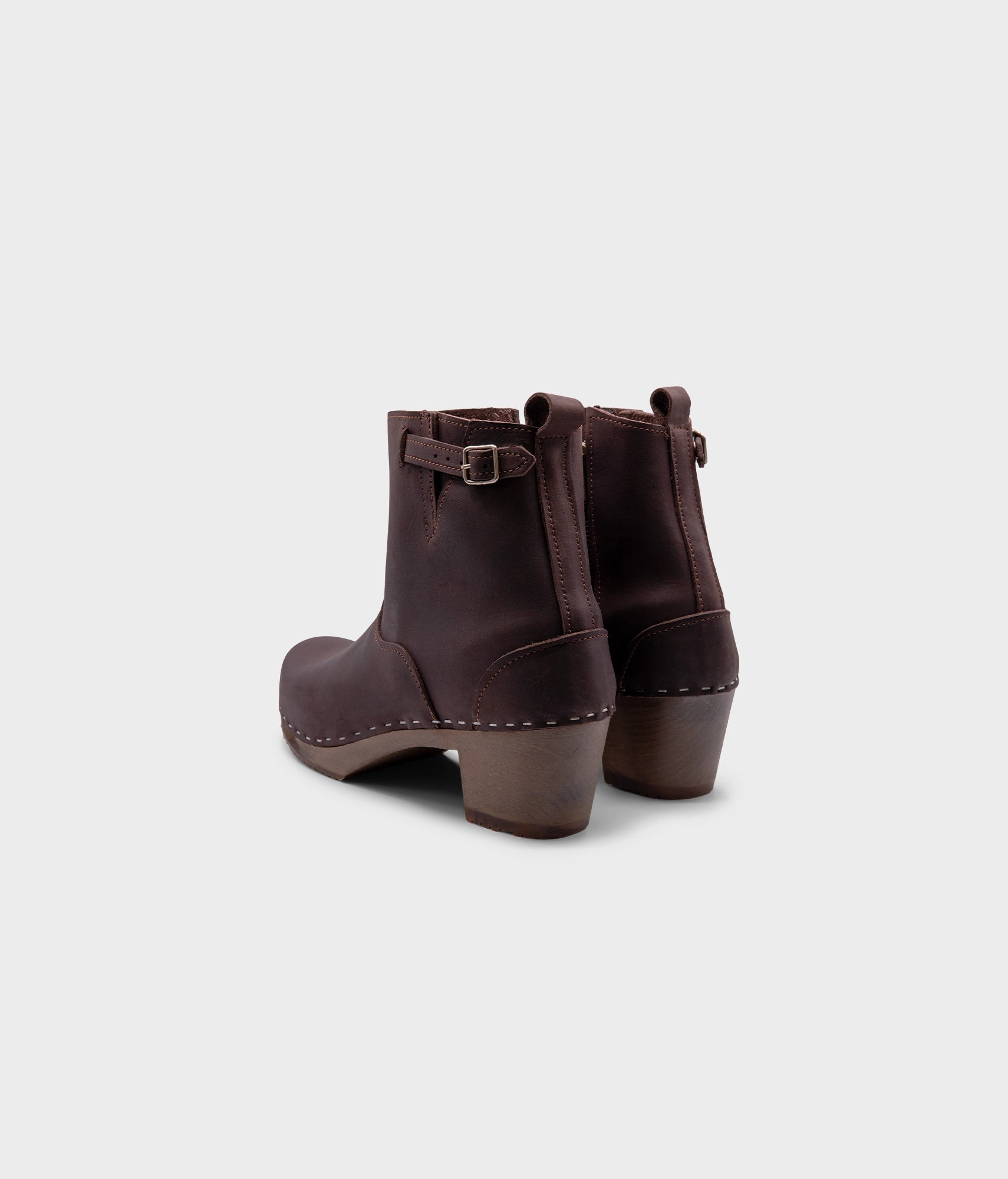 high-heeled clog boots in dark brown nubuck leather stapled on a dark wooden base with a silver buckle
