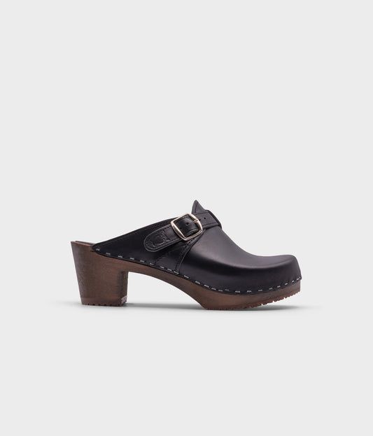 high heeled clog mules in black vegetable tanned leather with a silver buckle stapled on a dark wooden base