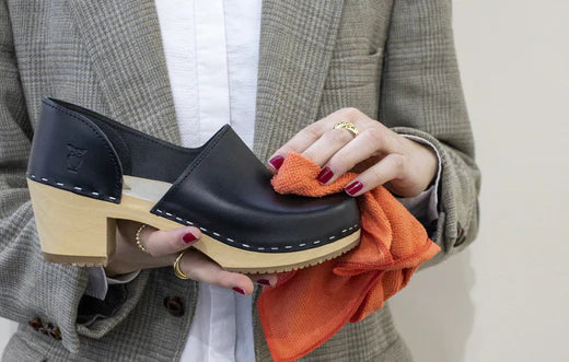 woman holding up a wooden clog in black shiny leather wiping it with a red cloth