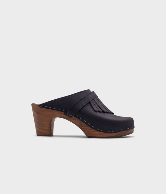 high rise clog mule in navy blue nubuck leather stapled on a dark wooden base with a leather strap with fringes