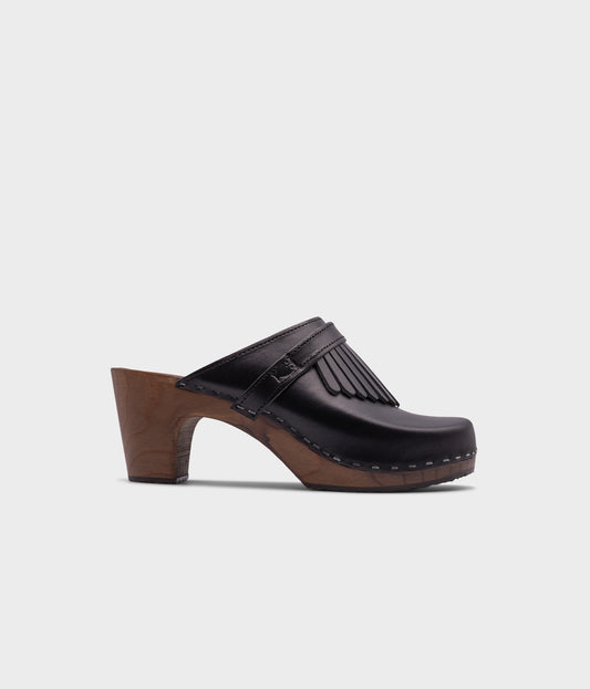high rise clog mule in black vegetable tanned leather stapled on a dark wooden base with a leather strap with fringes