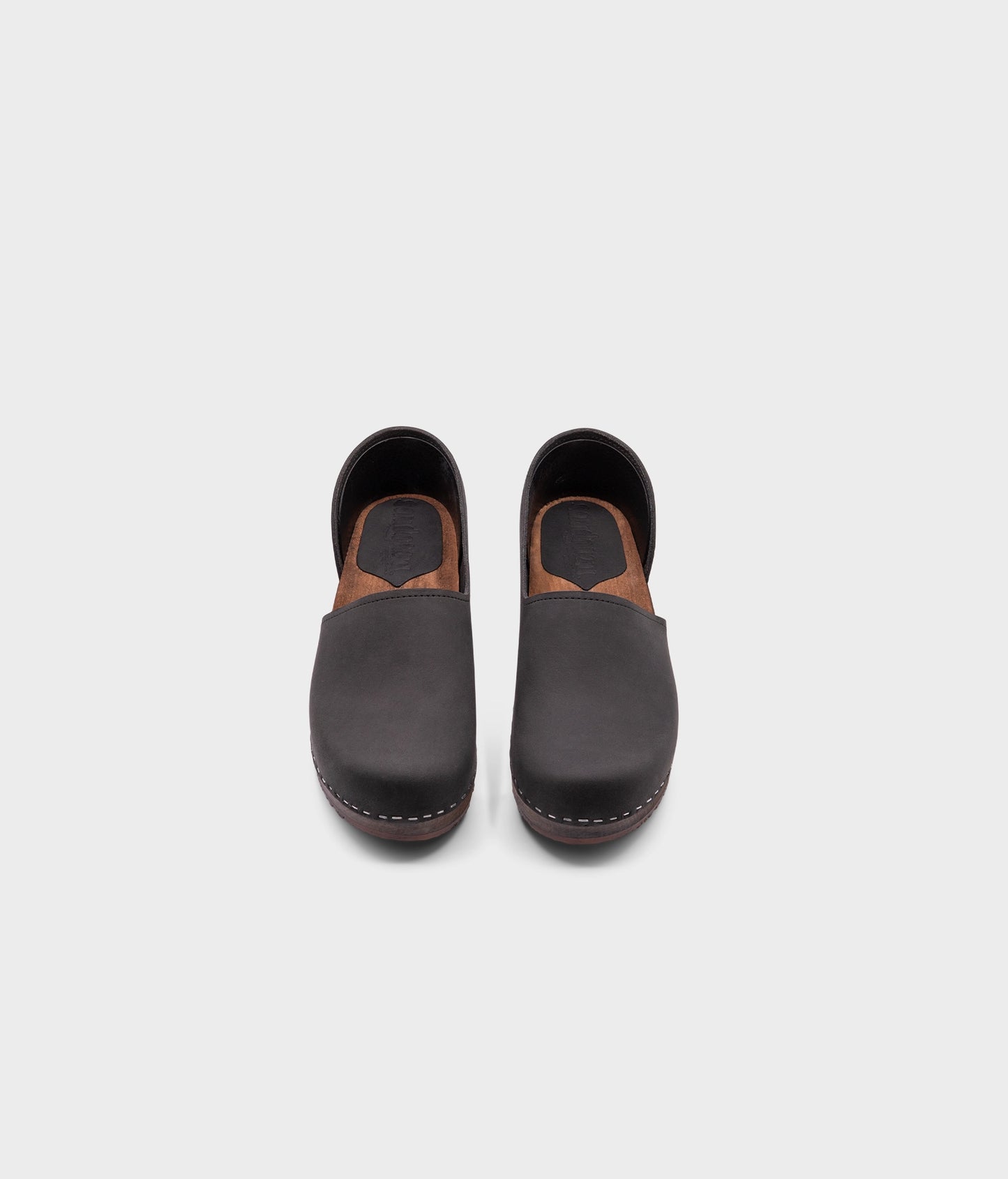 low heeled closed-back clogs in black nubuck leather stapled on a dark wooden base