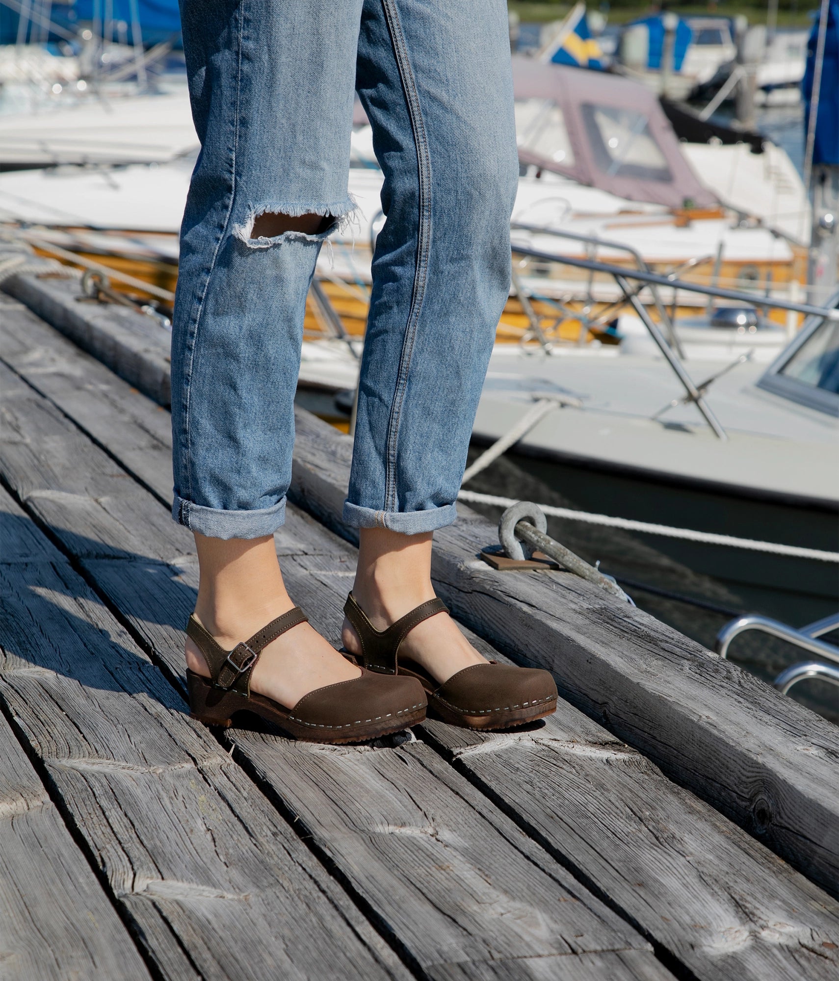 woman standing by a dock wearing ripped jeans and classic low heeled closed-toe sandal in dark brown nubuck leather stapled on a dark wooden base