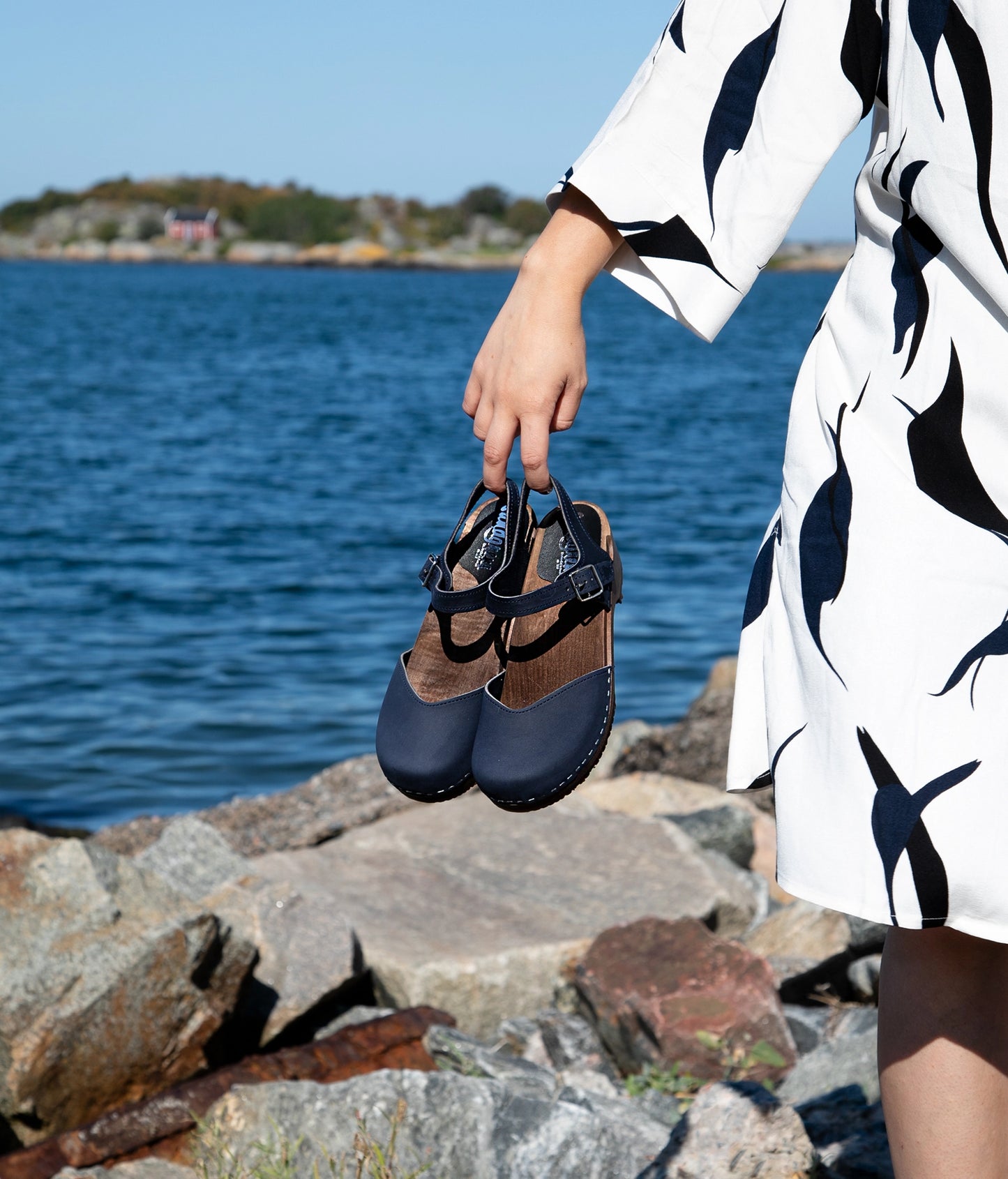 woman holding up a pair of classic low heeled closed-toe sandal in navy blue nubuck leather stapled on a dark wooden base by the ocean