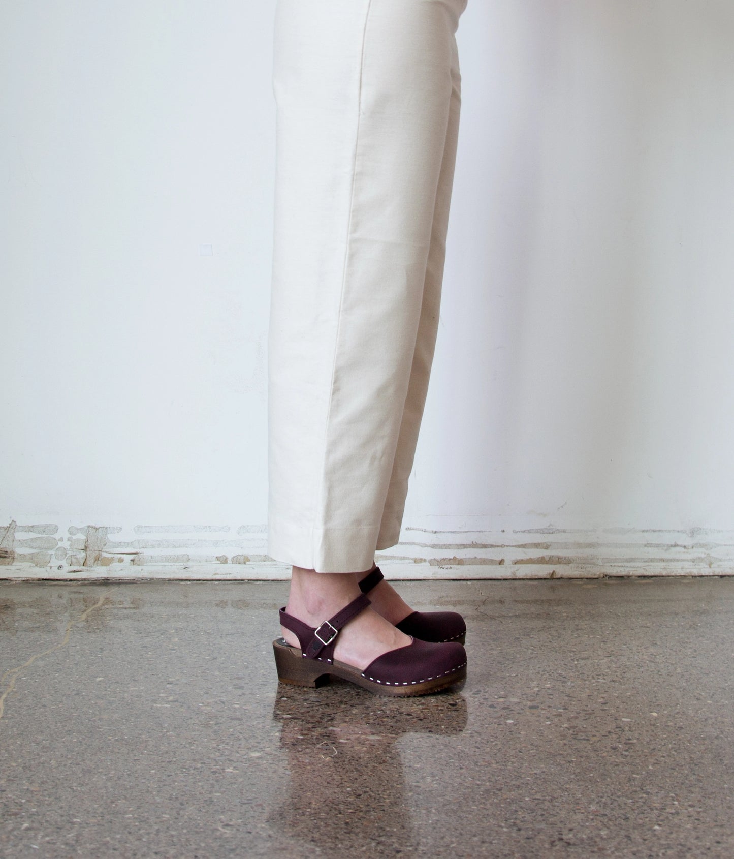 woman wearing white linen pants and classic low heeled closed-toe sandal in purple plum nubuck leather stapled on a dark wooden base