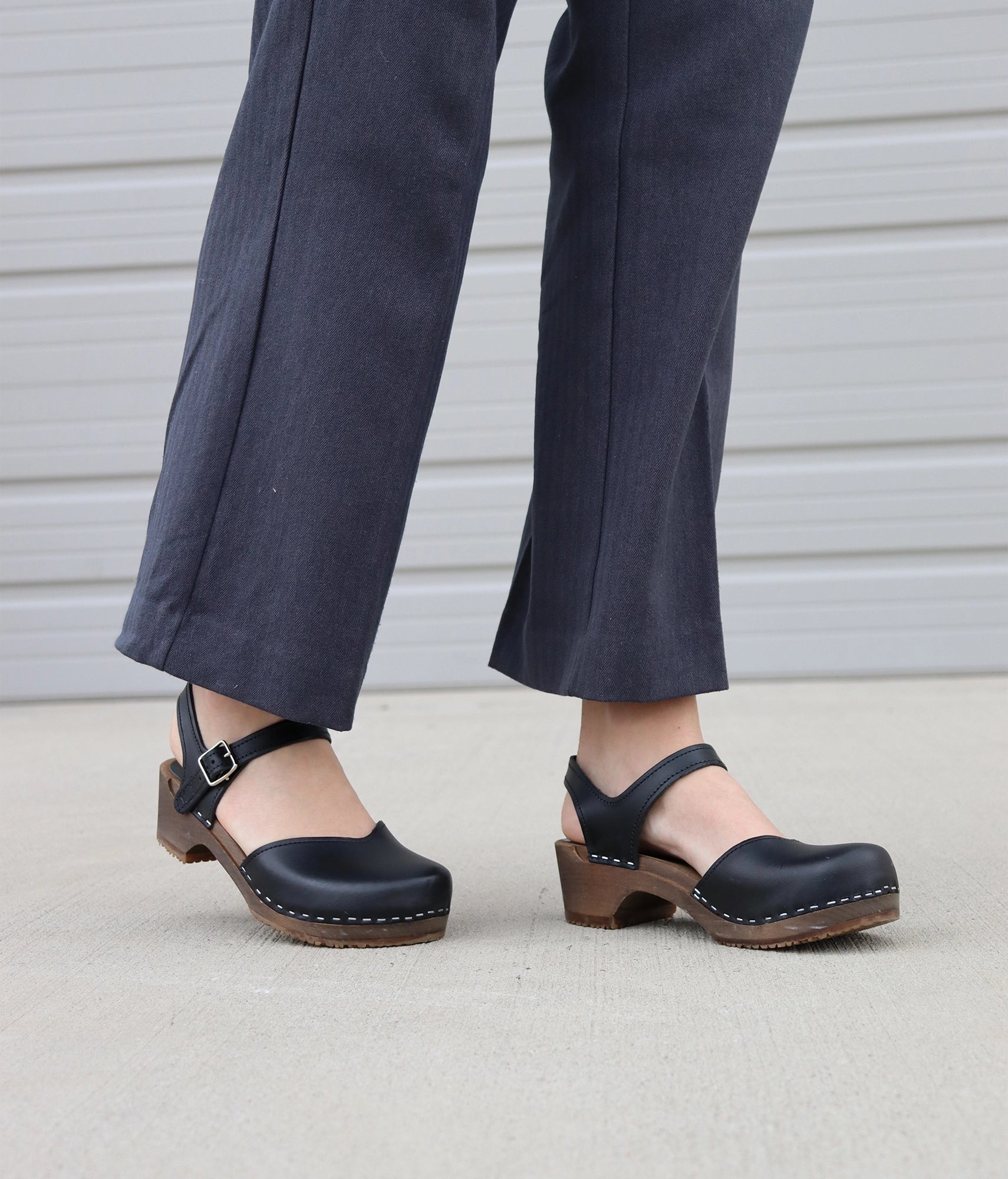 woman wearing grey dress pants and classic low heeled closed-toe sandal in black vegetable tanned leather stapled on a dark wooden base