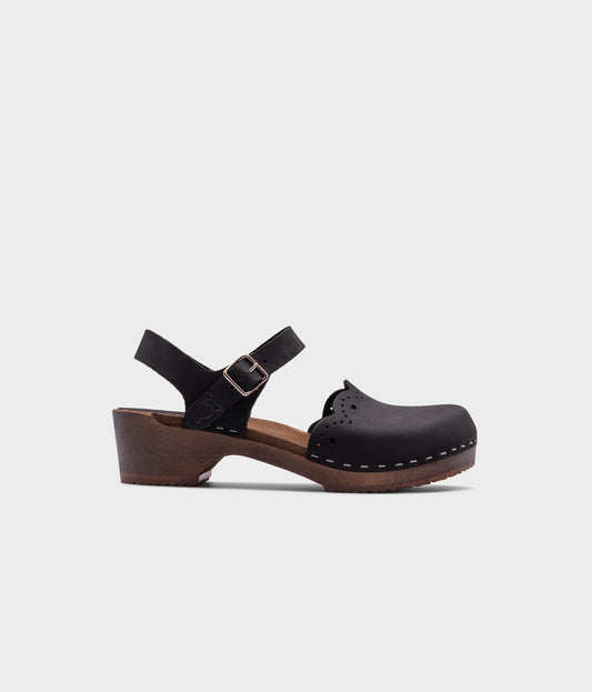 low heeled closed-toe clog sandals in black nubuck leather with a wavy leather cutout stapled on a dark wooden base