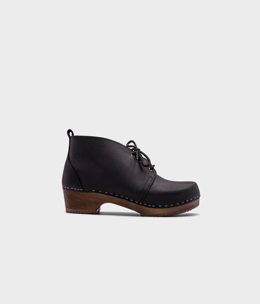 low heeled mens chukka clog boots in black nubuck leather stapled on a dark wooden base with black laces