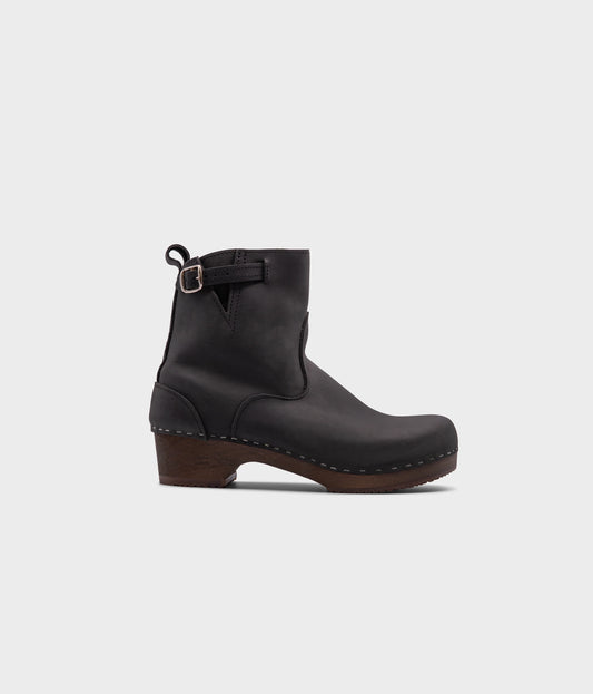 low heeled clog boots in black nubuck leather with a mid-shaft and silver buckle, stapled on a dark wooden base