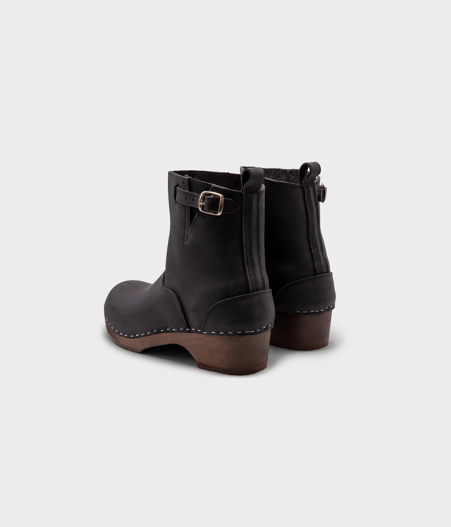 low heeled clog boots in black nubuck leather with a mid-shaft and silver buckle, stapled on a dark wooden base