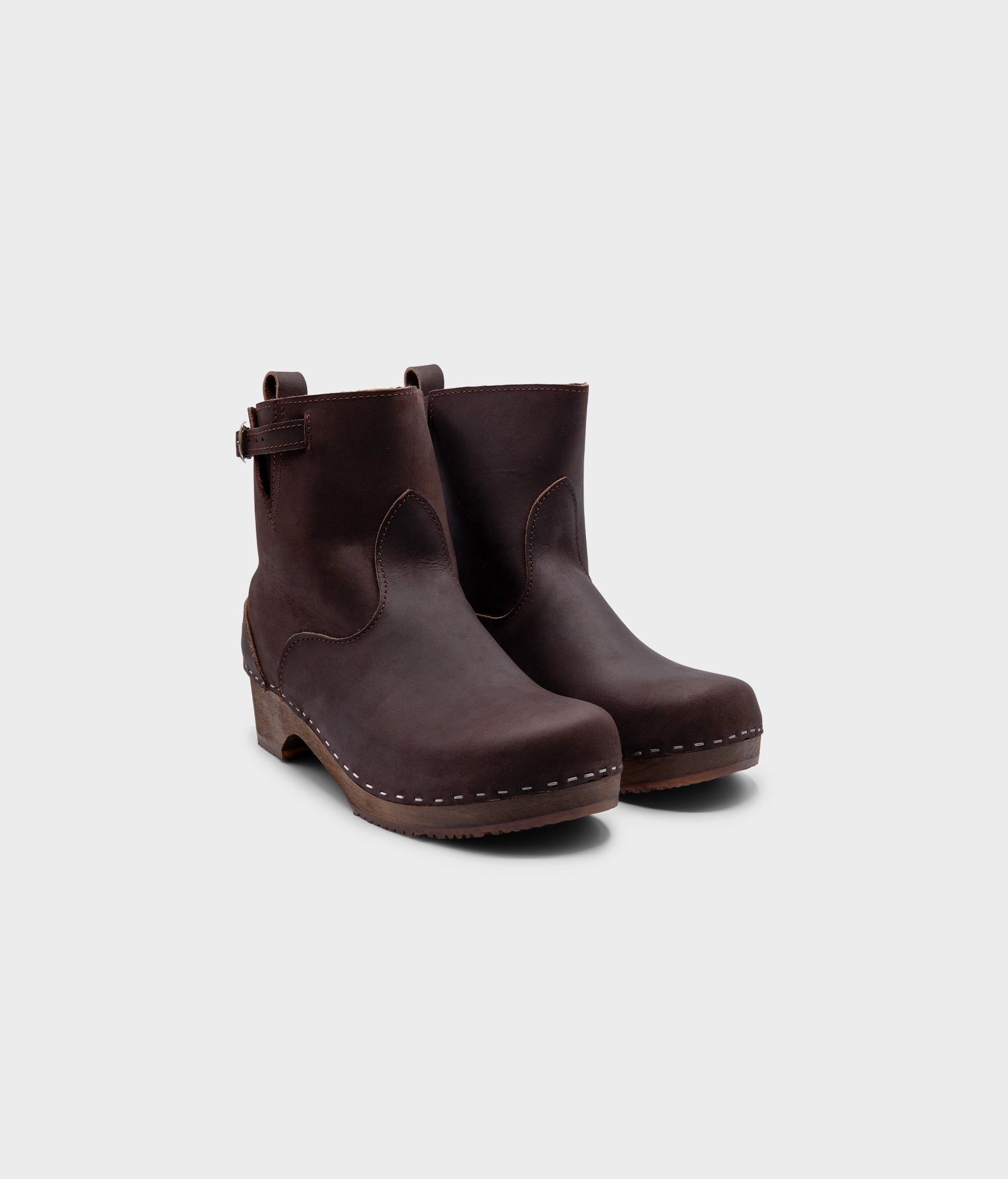 low heeled clog boots in dark brown nubuck leather with a mid-shaft and silver buckle, stapled on a dark wooden base