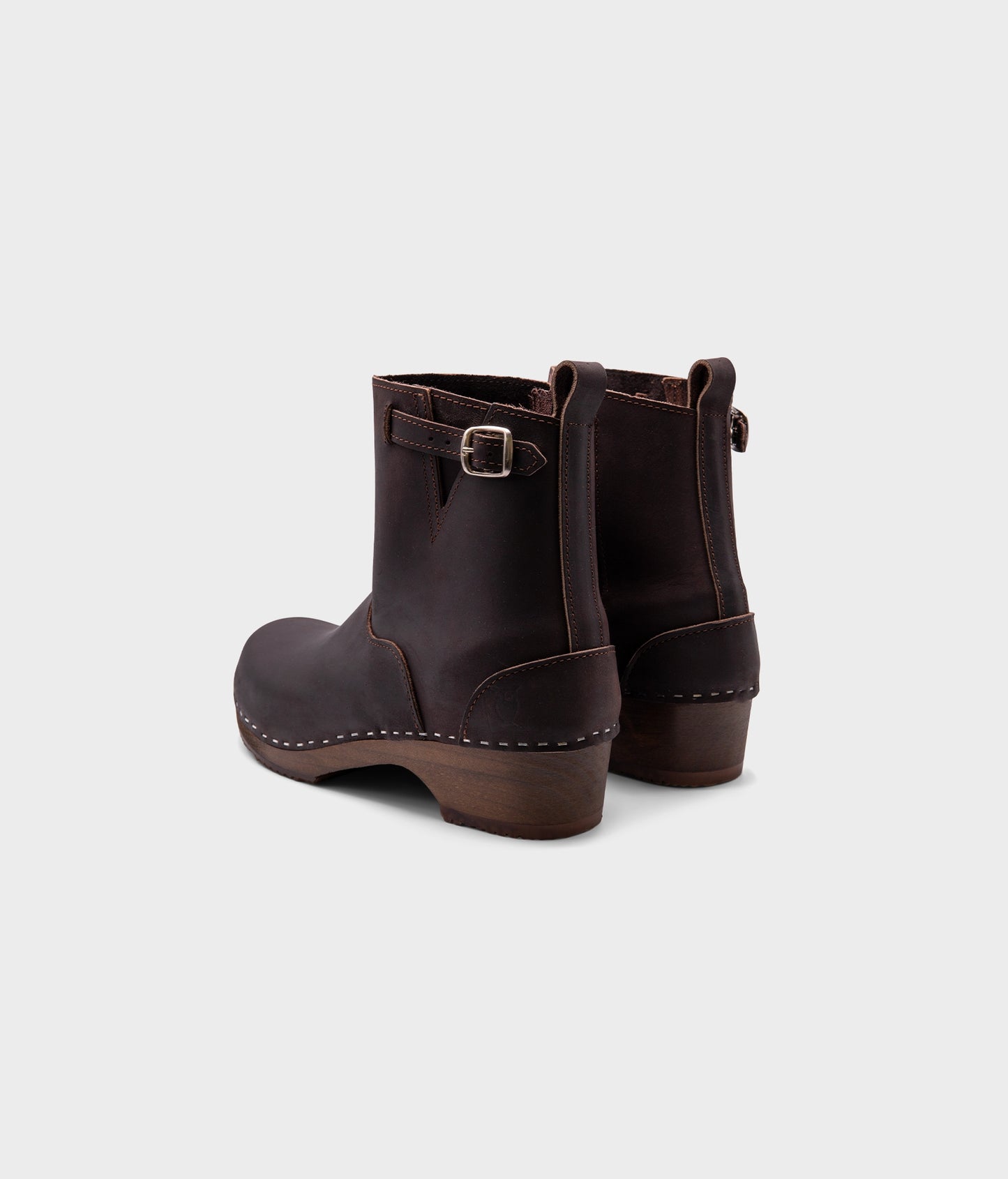 low heeled clog boots in dark brown nubuck leather with a mid-shaft and silver buckle, stapled on a dark wooden base