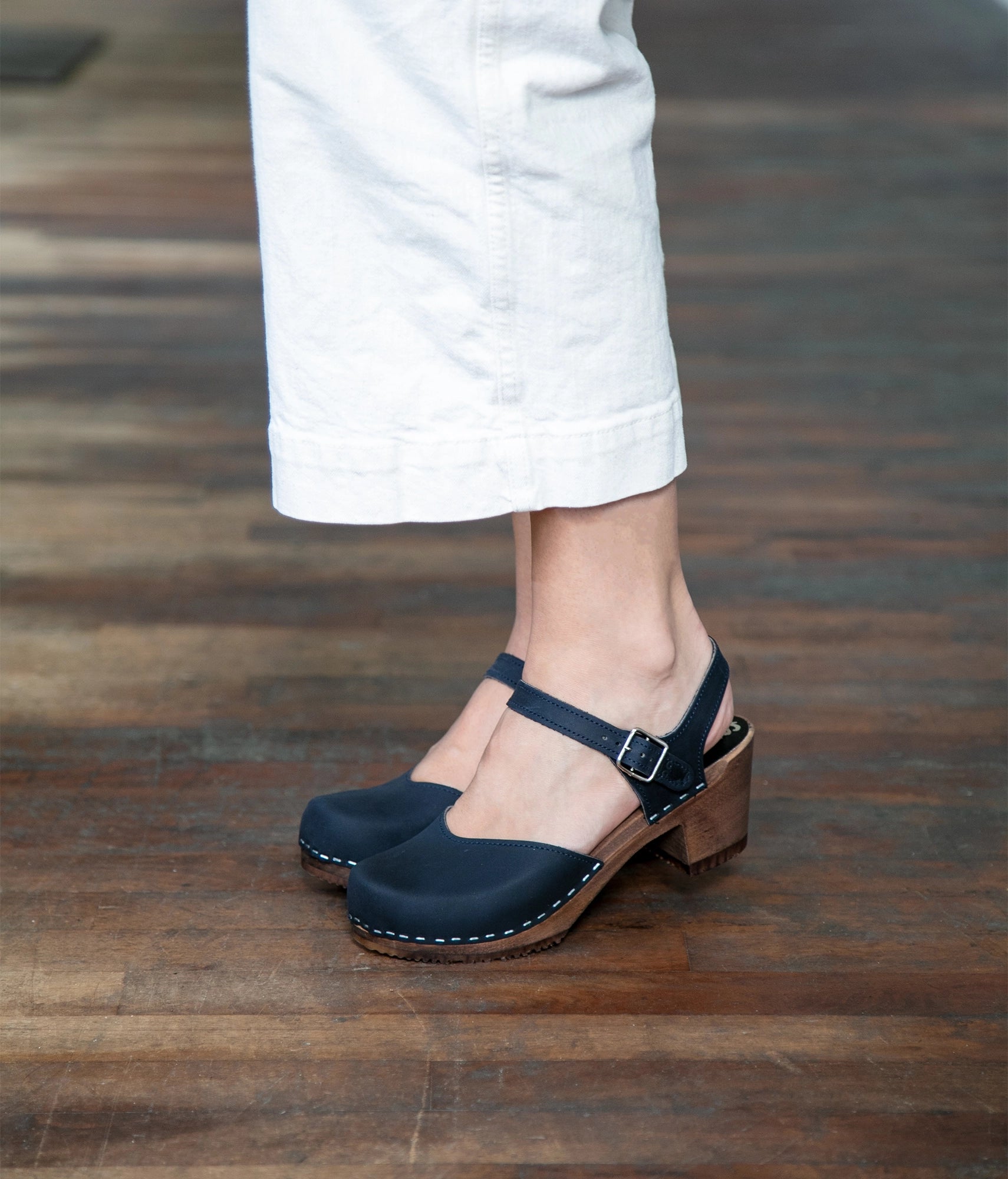 woman wearing white jeans and high heeled closed-toe clog sandal in navy blue nubuck leather stapled on a dark wooden base