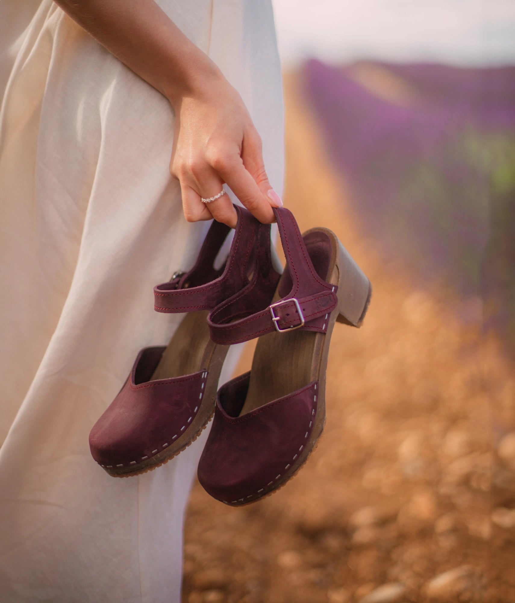woman holding a pair of high heeled closed-toe clog sandal in purple plum nubuck leather stapled on a dark wooden base