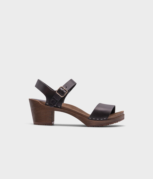high-heeled open-toe clog sandal in black vegetable tanned leather stapled on a dark wooden base