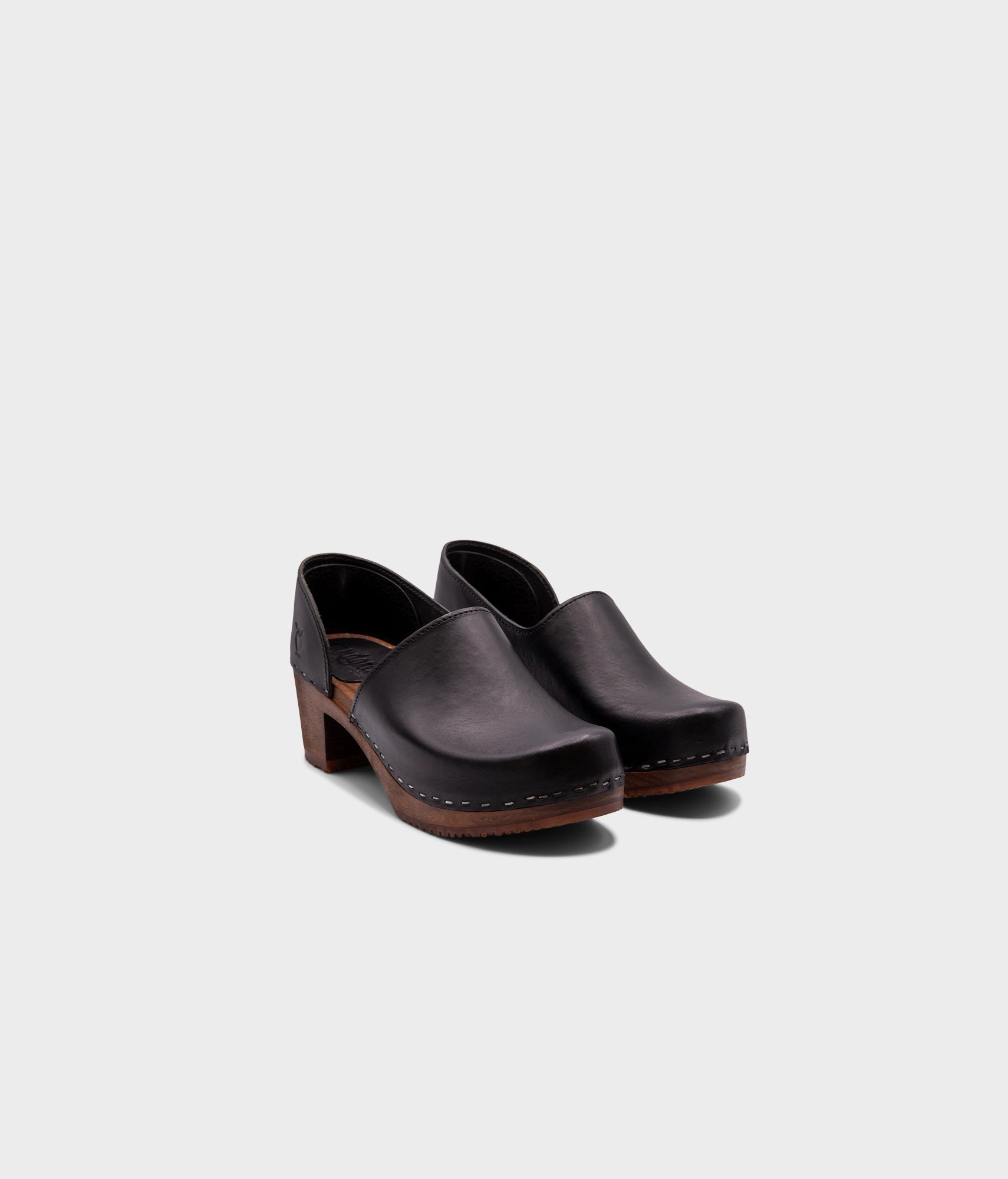 high heeled closed-back clogs in black vegetable tanned leather stapled on a dark wooden base