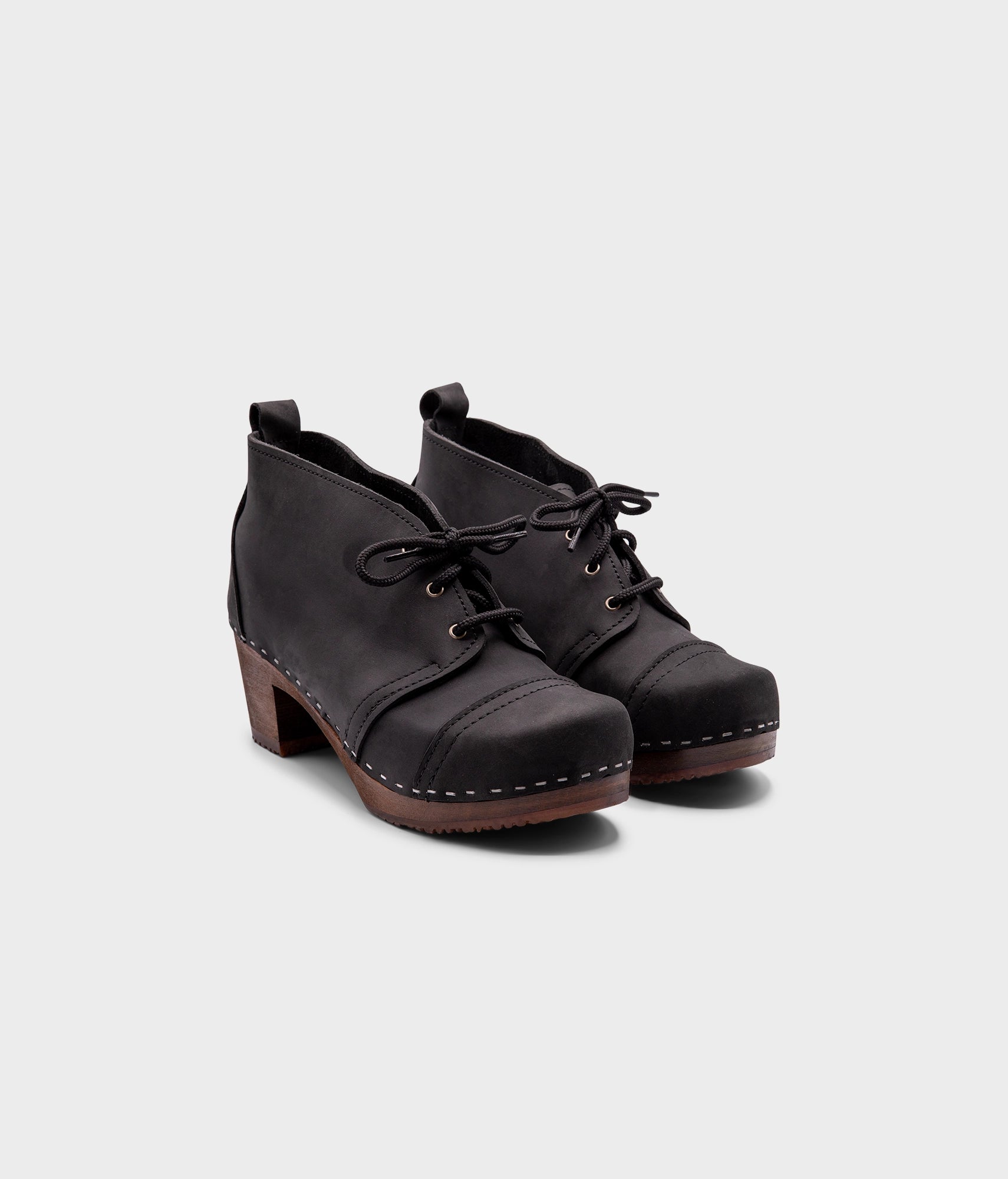 high heeled chukka clog boots in black nubuck leather stapled on a dark wooden base with black laces