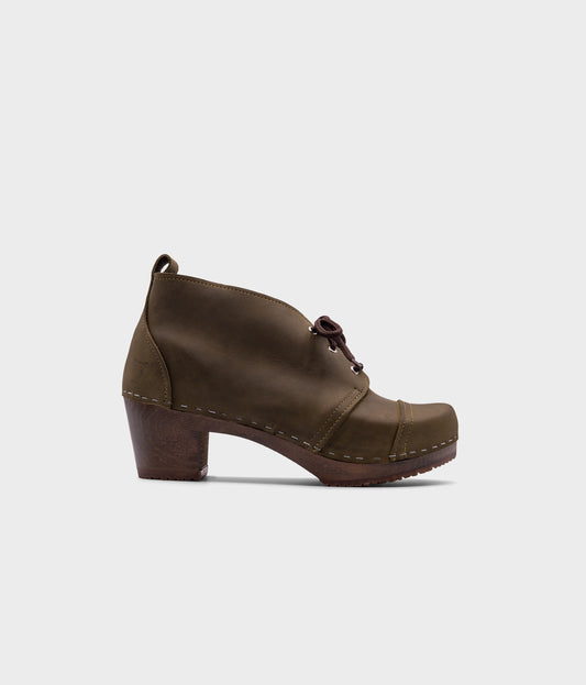 high heeled chukka clog boots in olive green nubuck leather stapled on a dark wooden base with brown laces