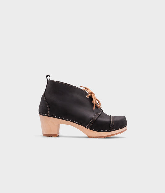 high heeled chukka clog boots in black nubuck leather stapled on a light wooden base with beige laces