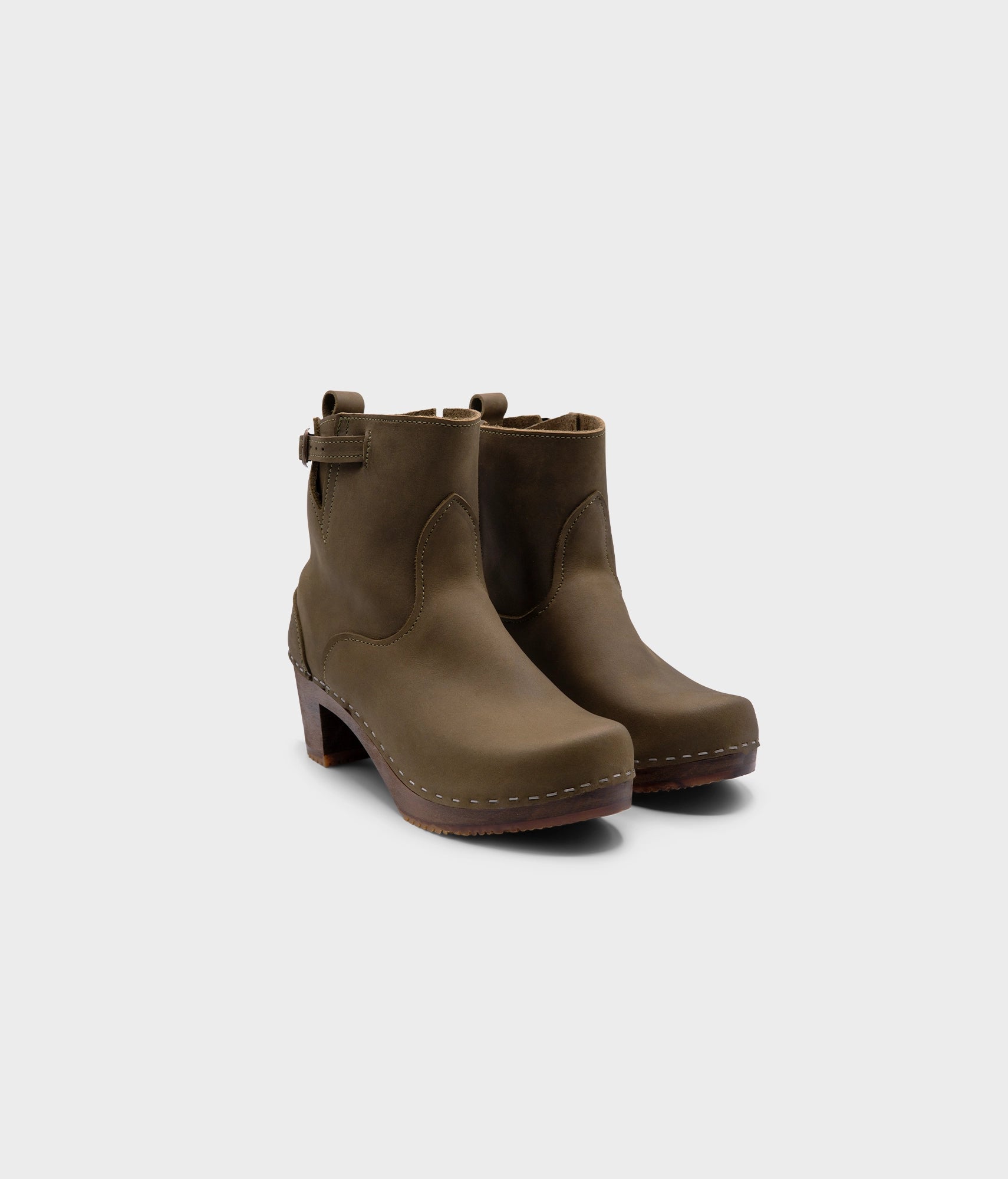 high-heeled clog boots in olive green nubuck leather stapled on a dark wooden base with a silver buckle