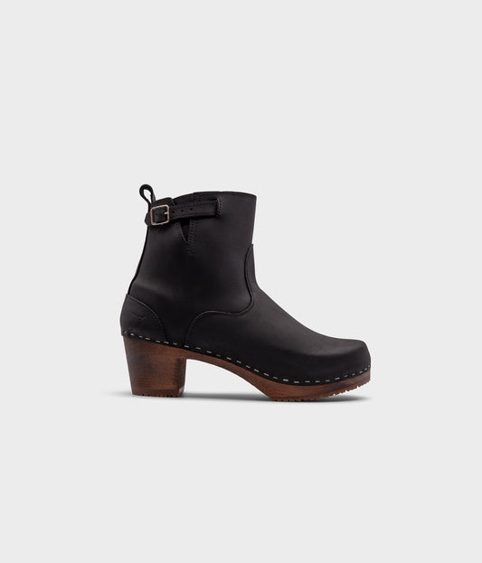 high-heeled clog boots in black nubuck leather stapled on a dark wooden base with a silver buckle