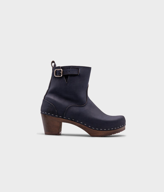 high-heeled clog boots in navy blue nubuck leather stapled on a dark wooden base with a silver buckle