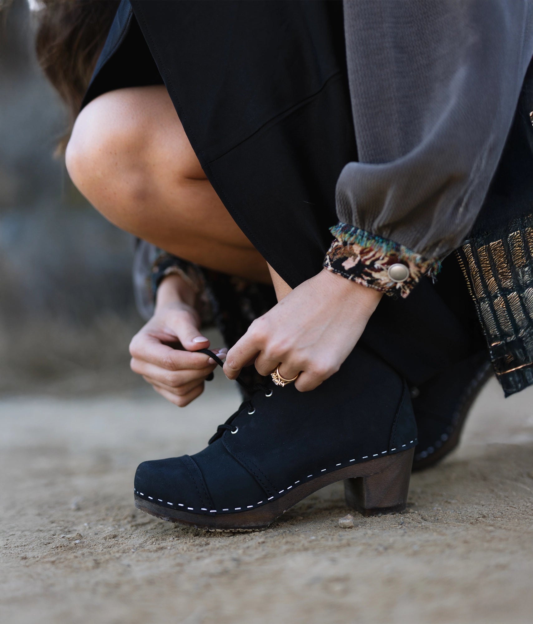 Person tying chukka can-toe clogs with a blurred background. High heeled chukka clog boots in black nubuck leather stapled on a dark wooden base with black laces