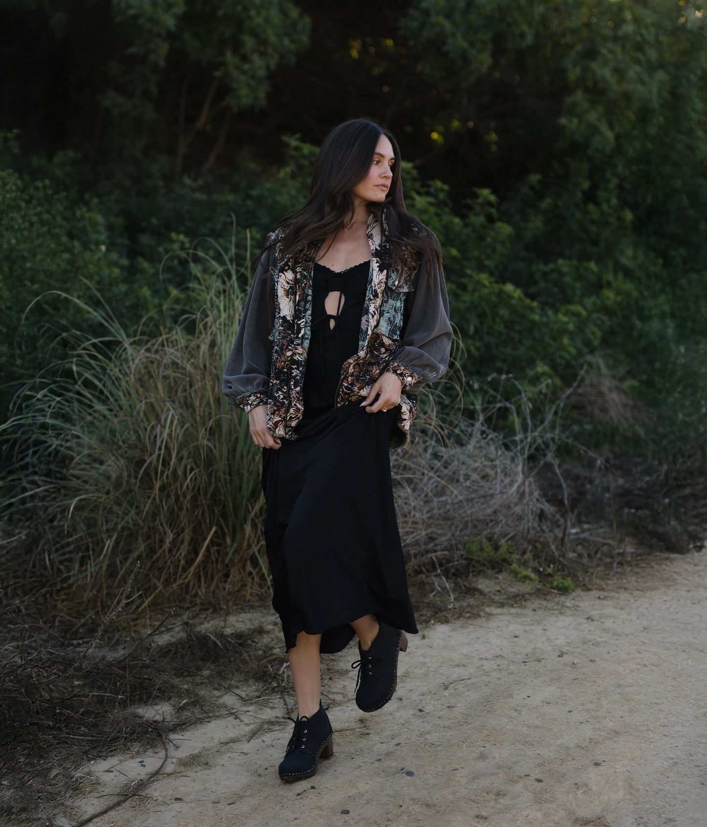 Woman walking outdoors in a natural setting wearing a black dress. High heeled chukka clog boots in black nubuck leather stapled on a dark wooden base with black laces