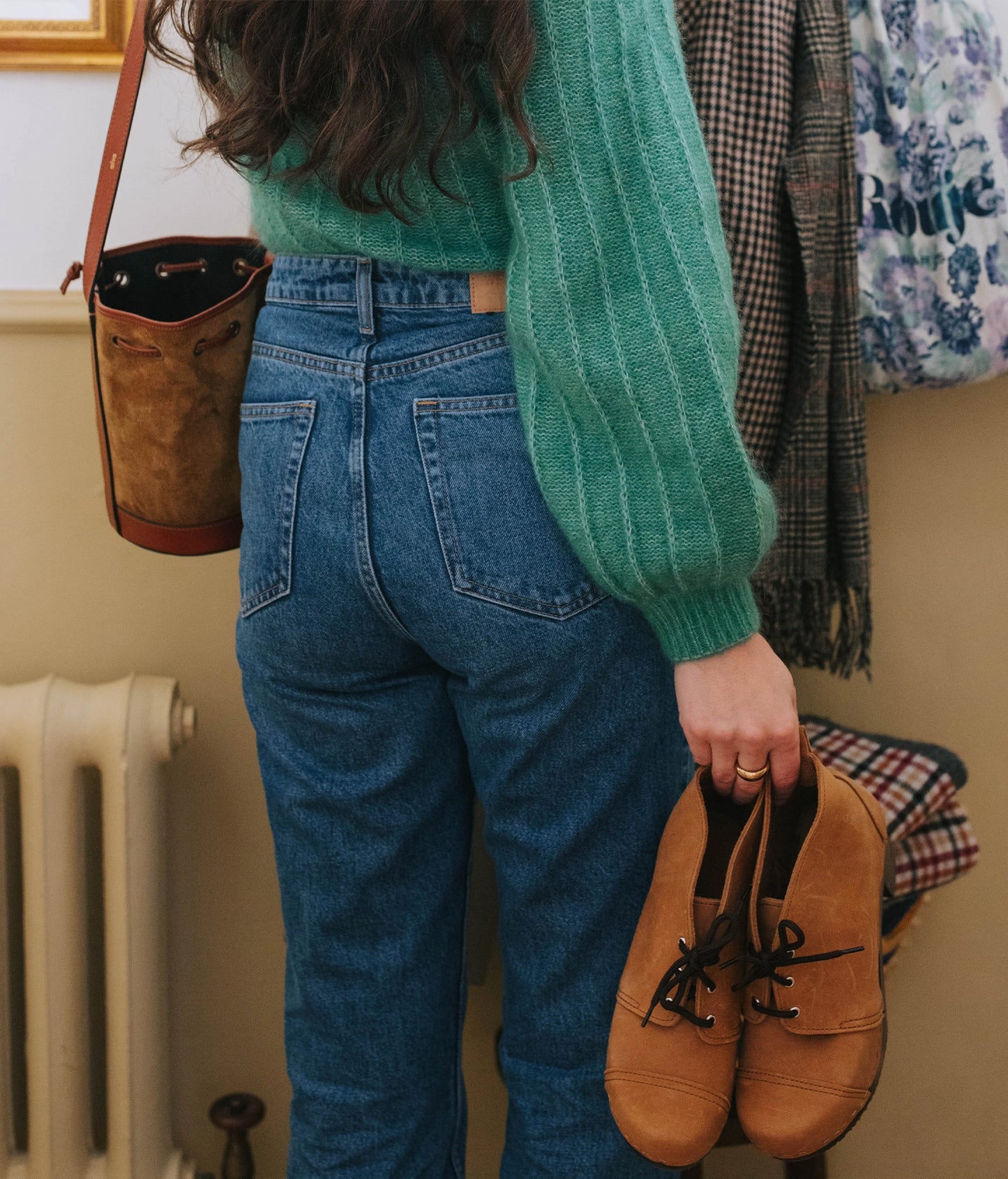 Person wearing a green sweater and blue jeans holding dextertan chukka cap-toe boots, standing in front of a radiator and coat rack.