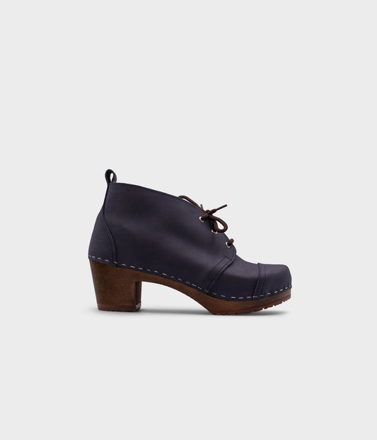 high heeled chukka clog boots in navy blue nubuck leather stapled on a dark wooden base with brown laces