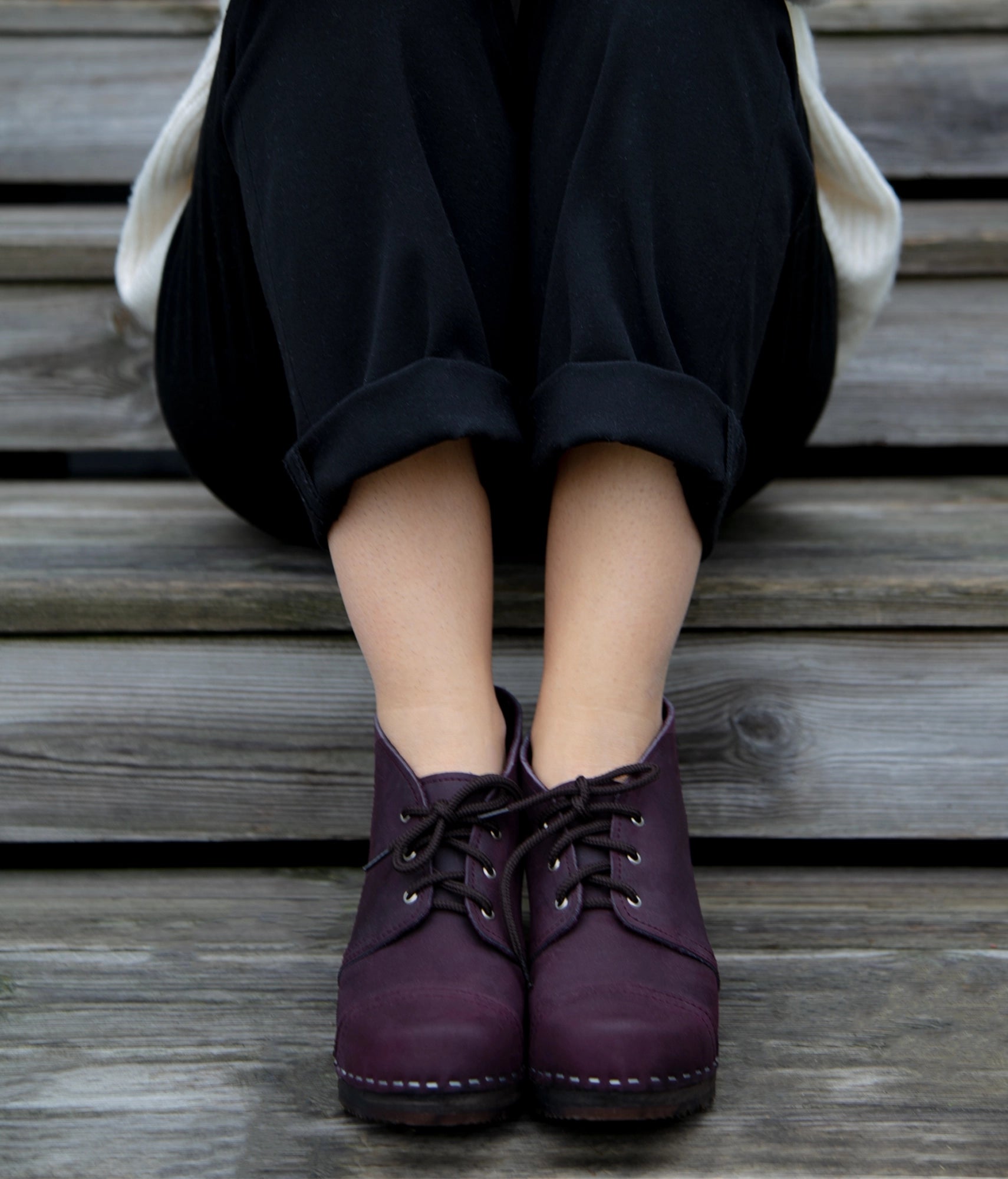 Person wearing plums colored Chukka cap-toe clogs sitting on wooden steps. High heeled chukka clog boots in purple plum nubuck leather stapled on a dark wooden base with brown laces