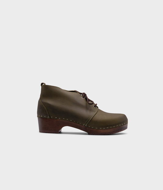 low heeled mens chukka clog boots in olive green nubuck leather stapled on a dark wooden base with brown laces