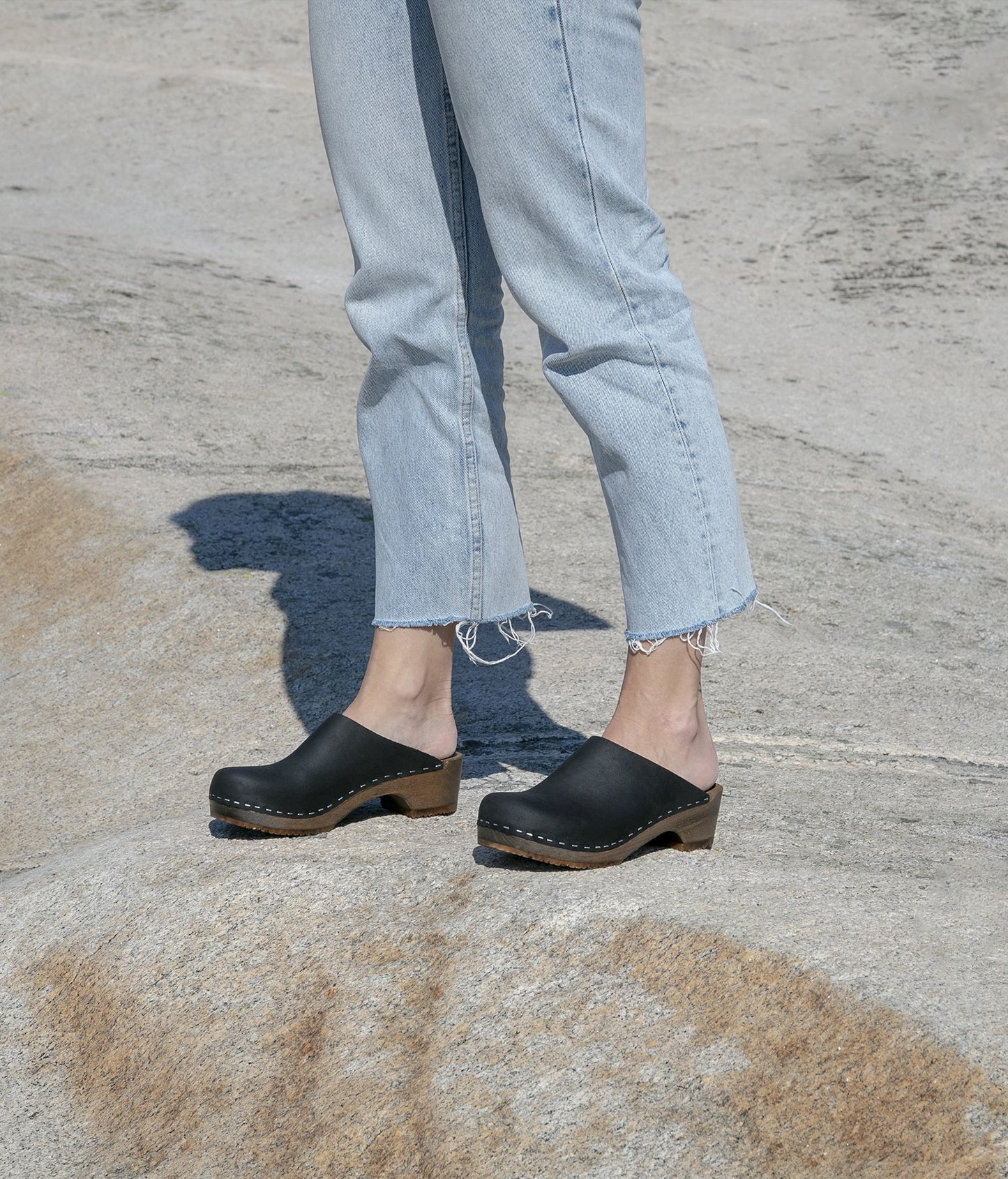 low heeled minimalistic clog mules in black nubuck leather stapled on a dark wooden base