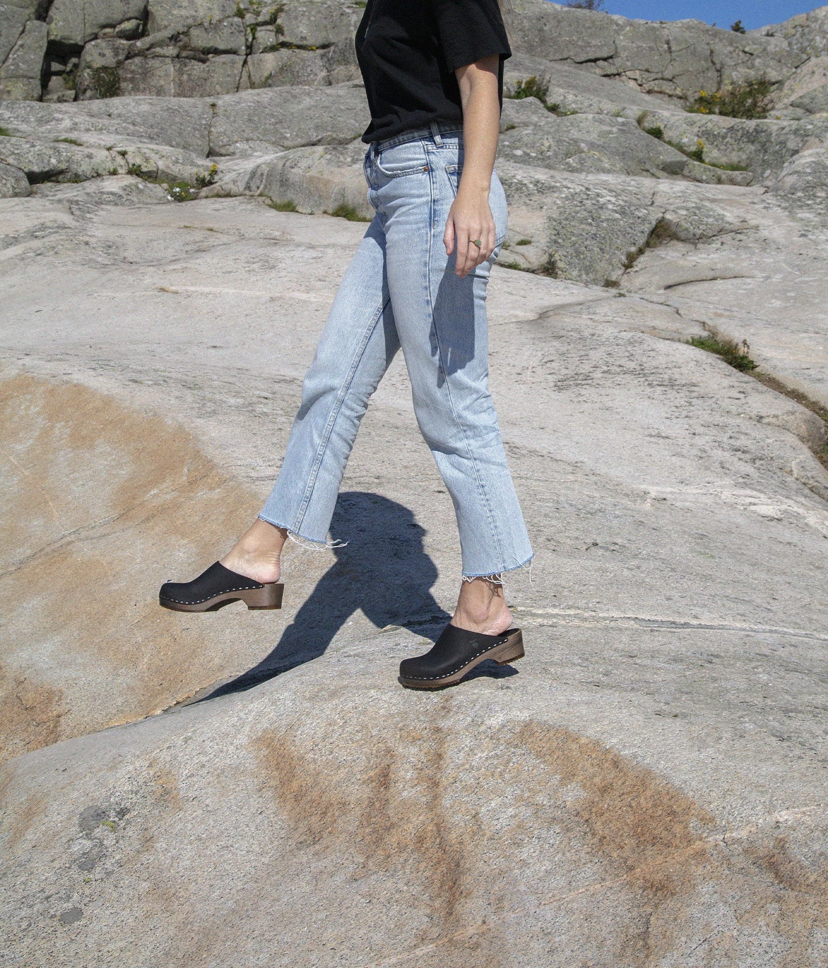 low heeled minimalistic clog mules in black nubuck leather stapled on a dark wooden base