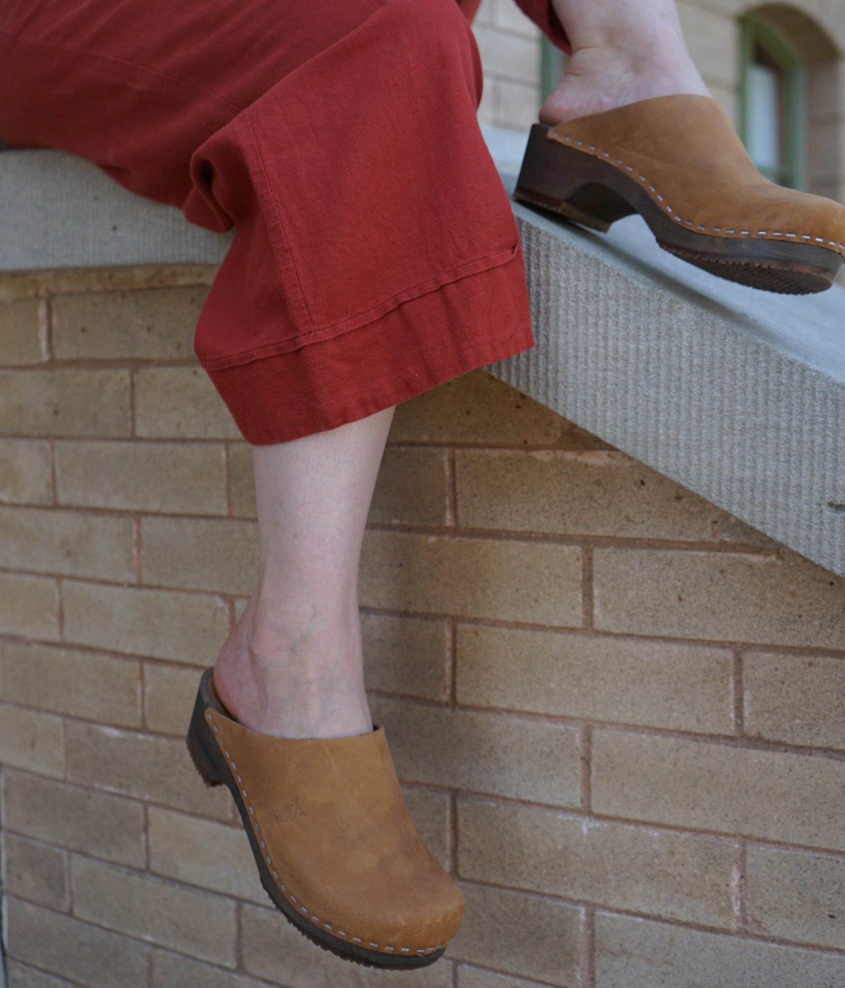 Person wearing Dexter tan colored clogs on a stone ledge with a brick wall background.