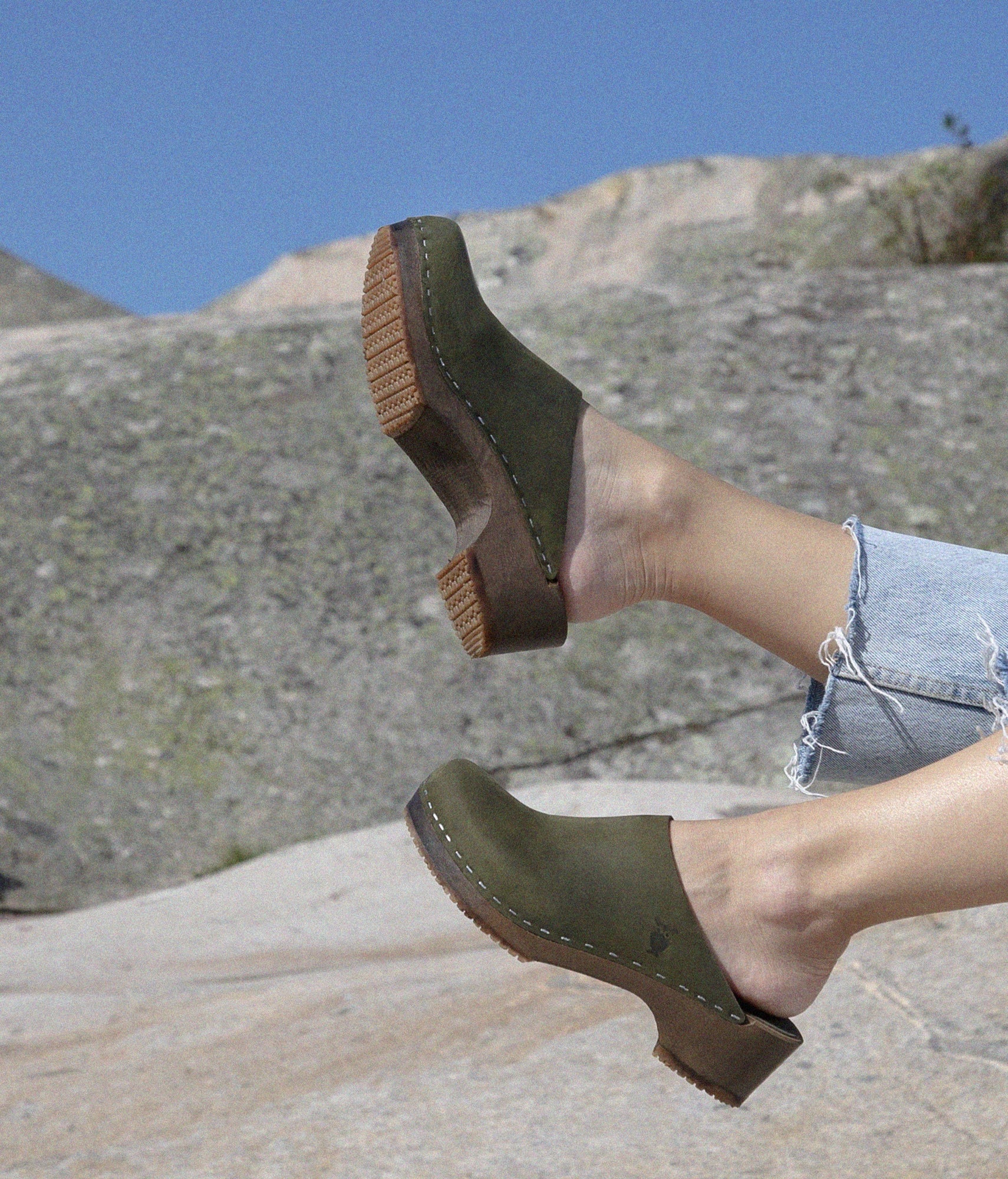 Person wearing olive green clogs with dark soles on a rocky surface. Low heeled minimalistic clog mules in olive green nubuck leather stapled on a dark wooden base