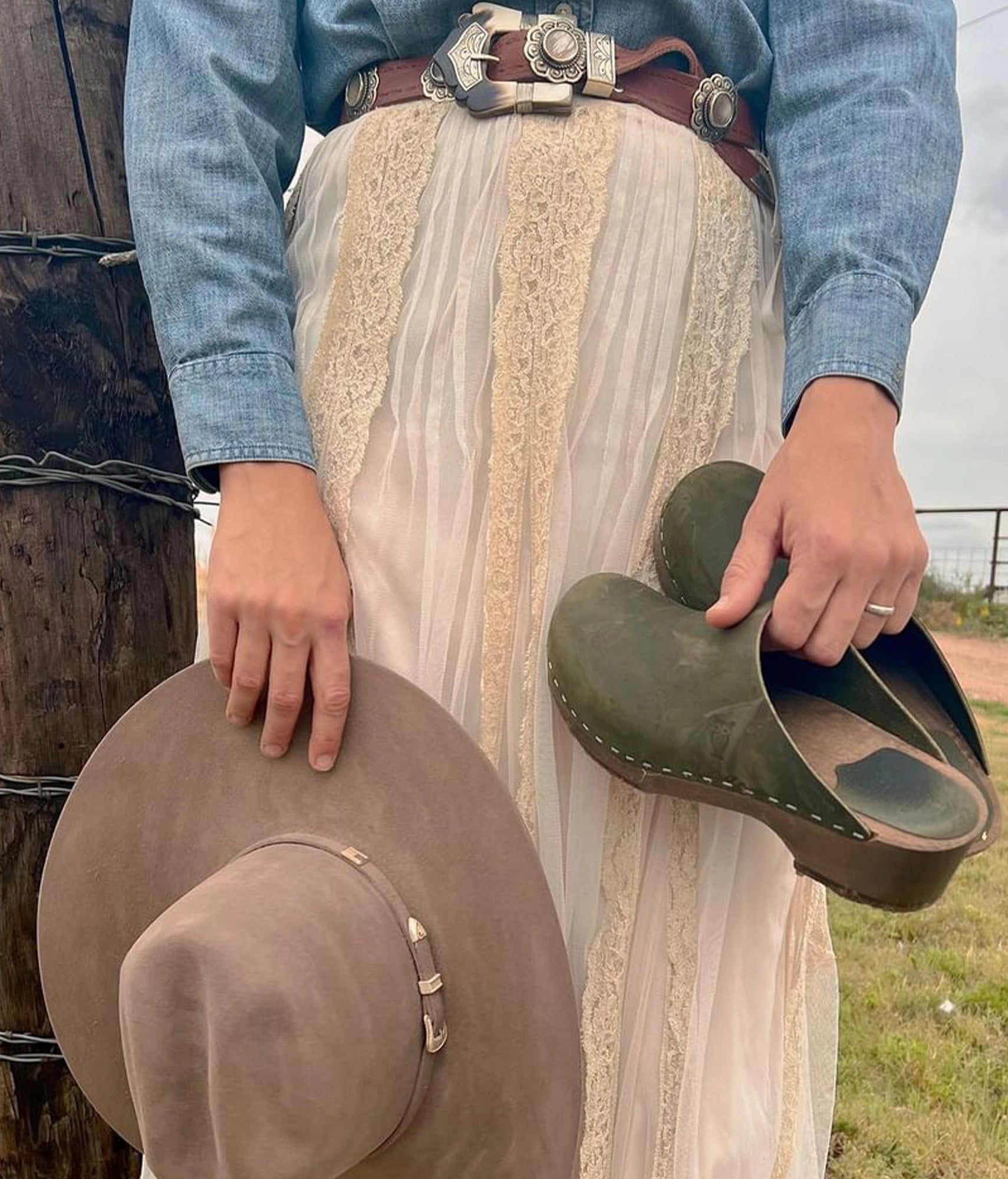 Person holding a cowboy hat and olive green clog mules outdoors. Low heeled minimalistic clog mules in olive green nubuck leather stapled on a dark wooden base