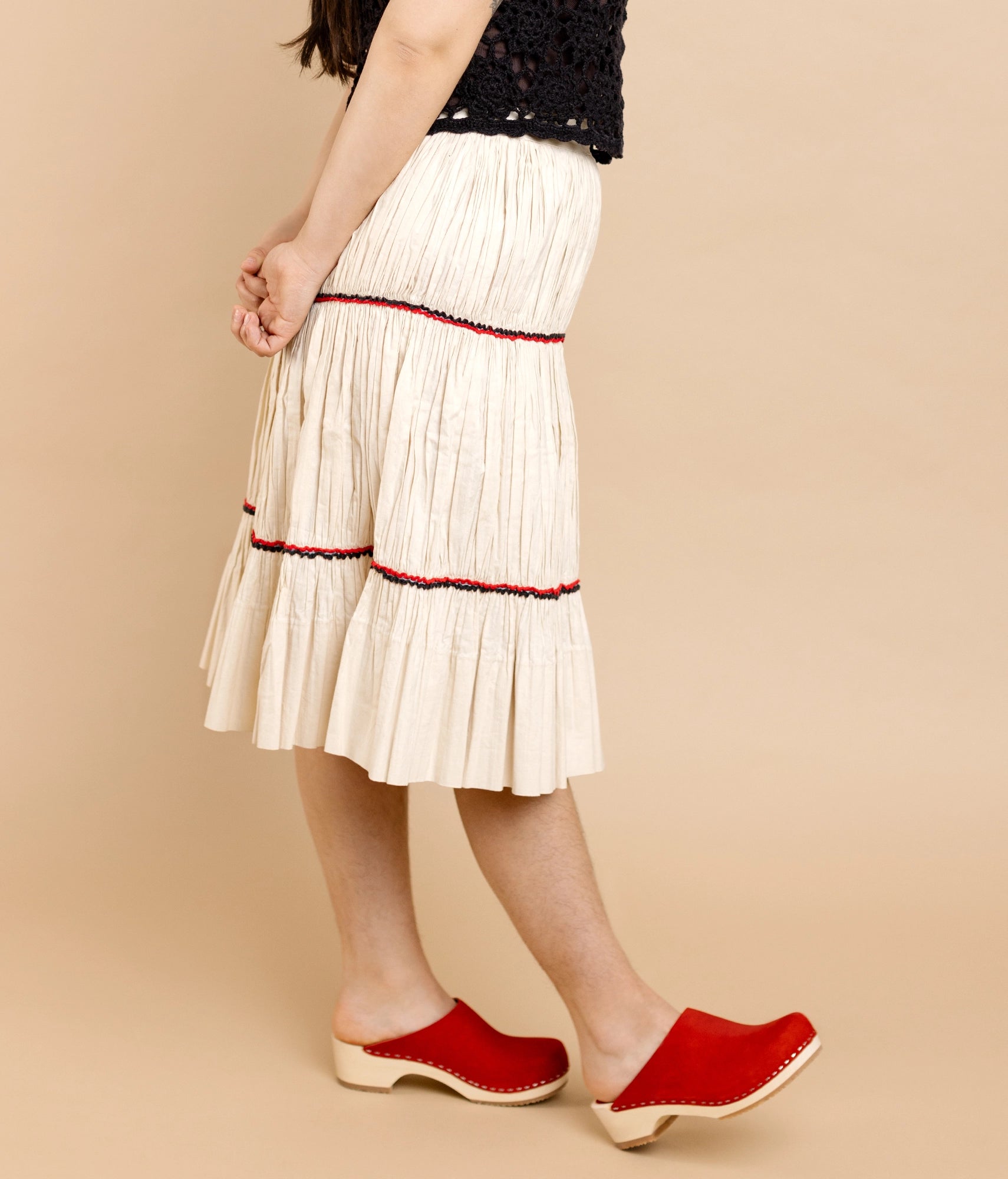 Person wearing a white pleated skirt with red trim and red clog mules on a beige background