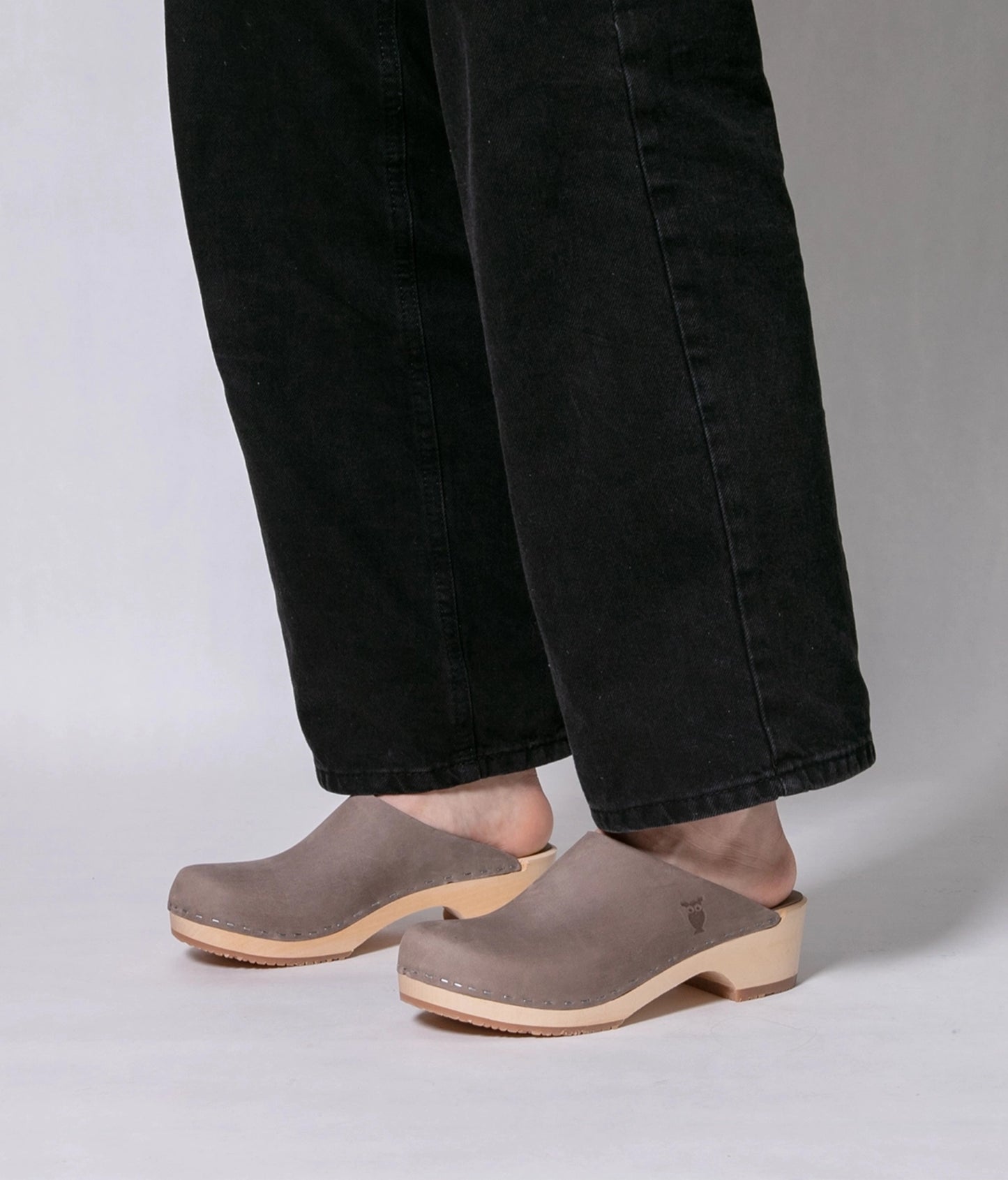 Person wearing Stone colored nubuck clogs with black jeans on a white background. low heeled minimalistic clog mules in stone grey nubuck leather stapled on a light wooden base