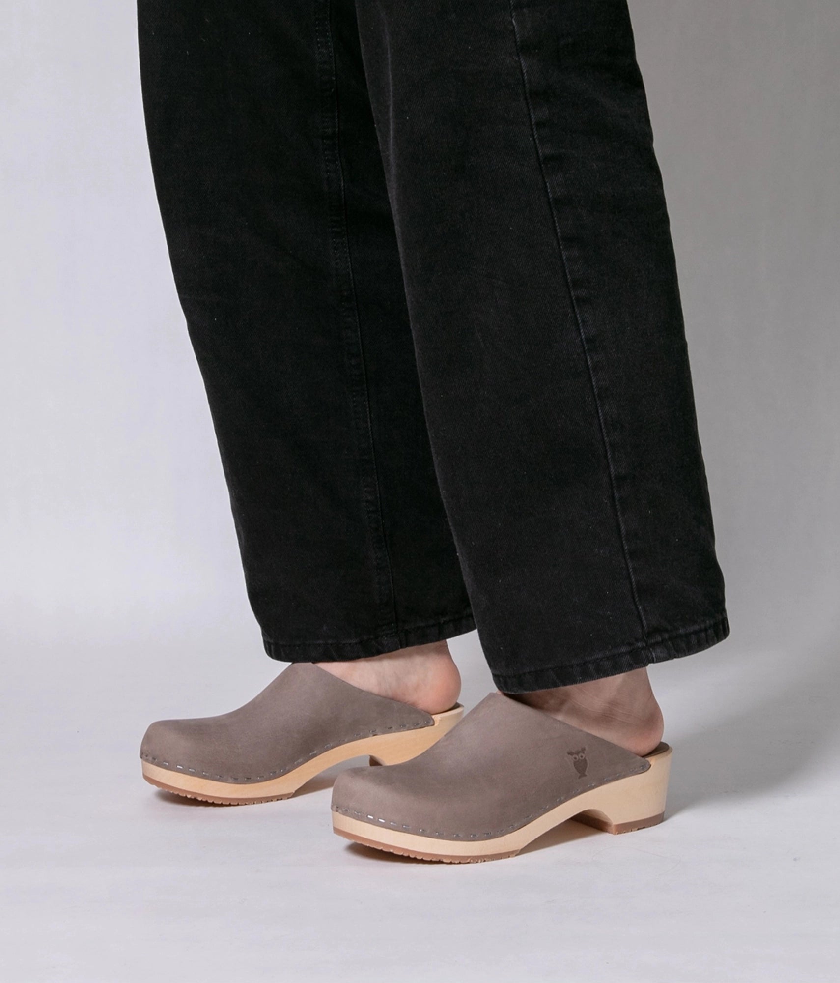 Person wearing Stone colored nubuck clogs with black jeans on a white background. low heeled minimalistic clog mules in stone grey nubuck leather stapled on a light wooden base