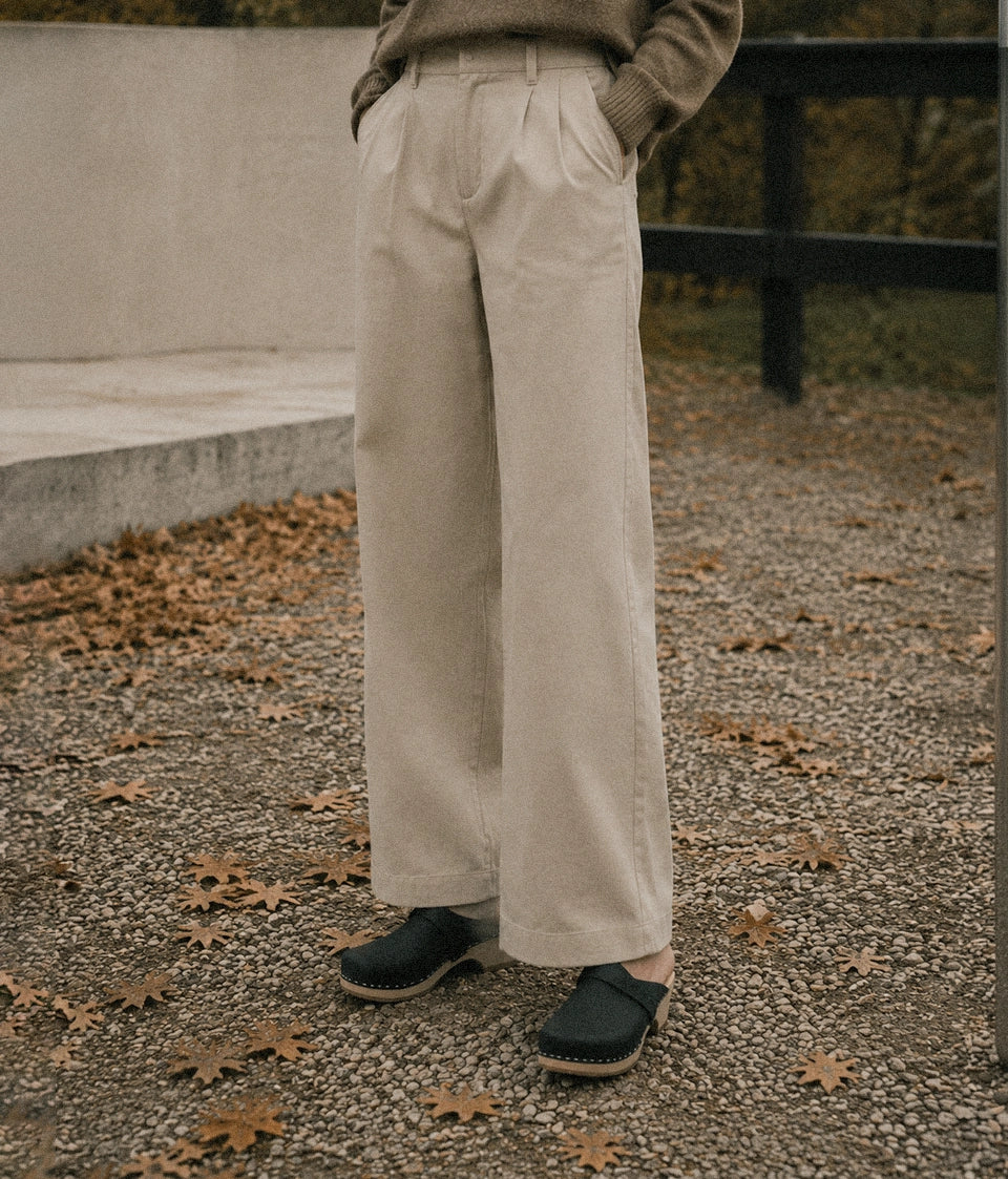 A person wearing light beige wide-leg trousers, a taupe knit sweater, and dark clog-style shoes, standing outdoors on a gravel path scattered with fallen leaves.
