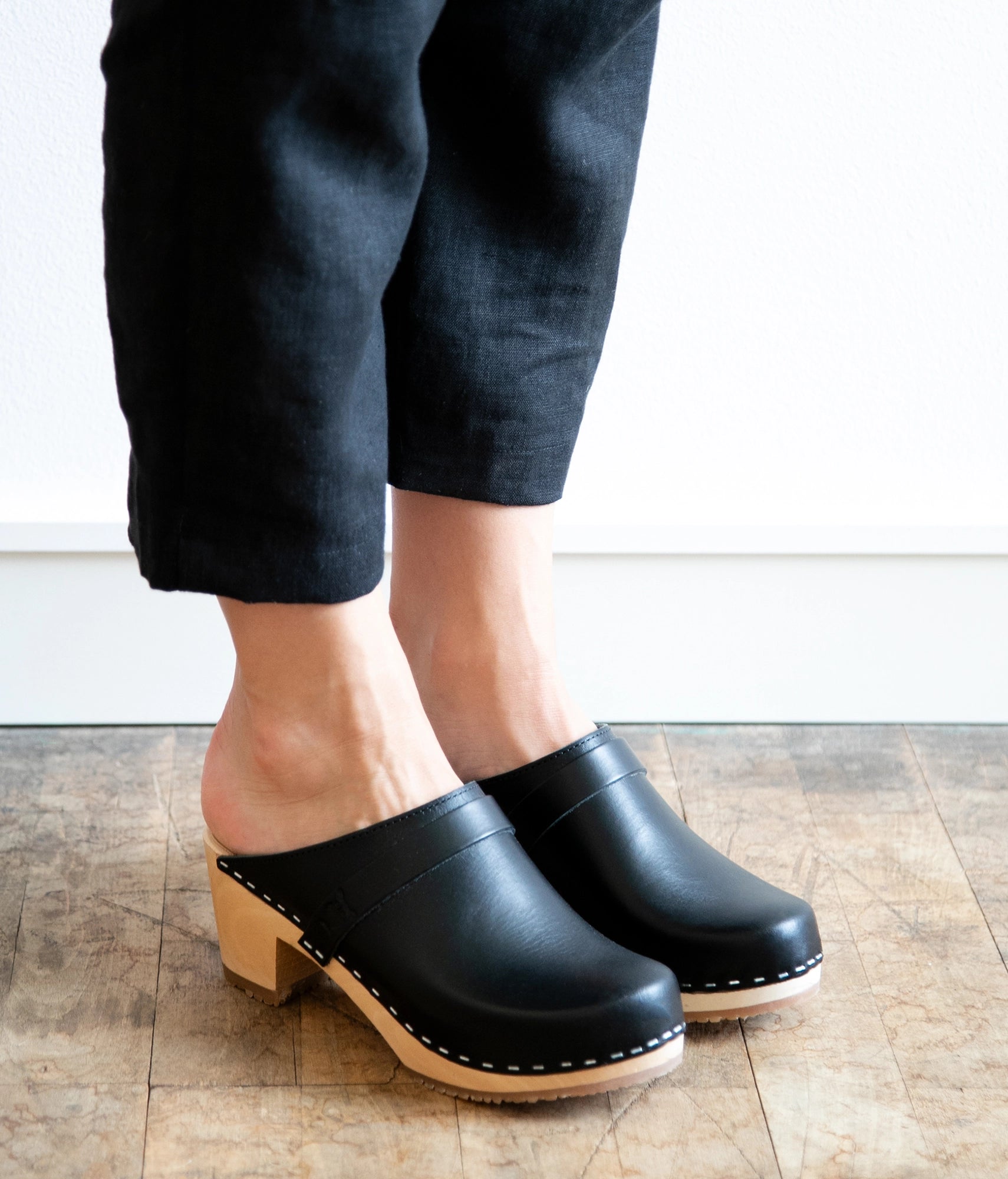 Black clogs worn with black pants on a wooden floor. classic high heeled clog mule in black vegetable tanned leather stapled on a light wooden base with a leather strap