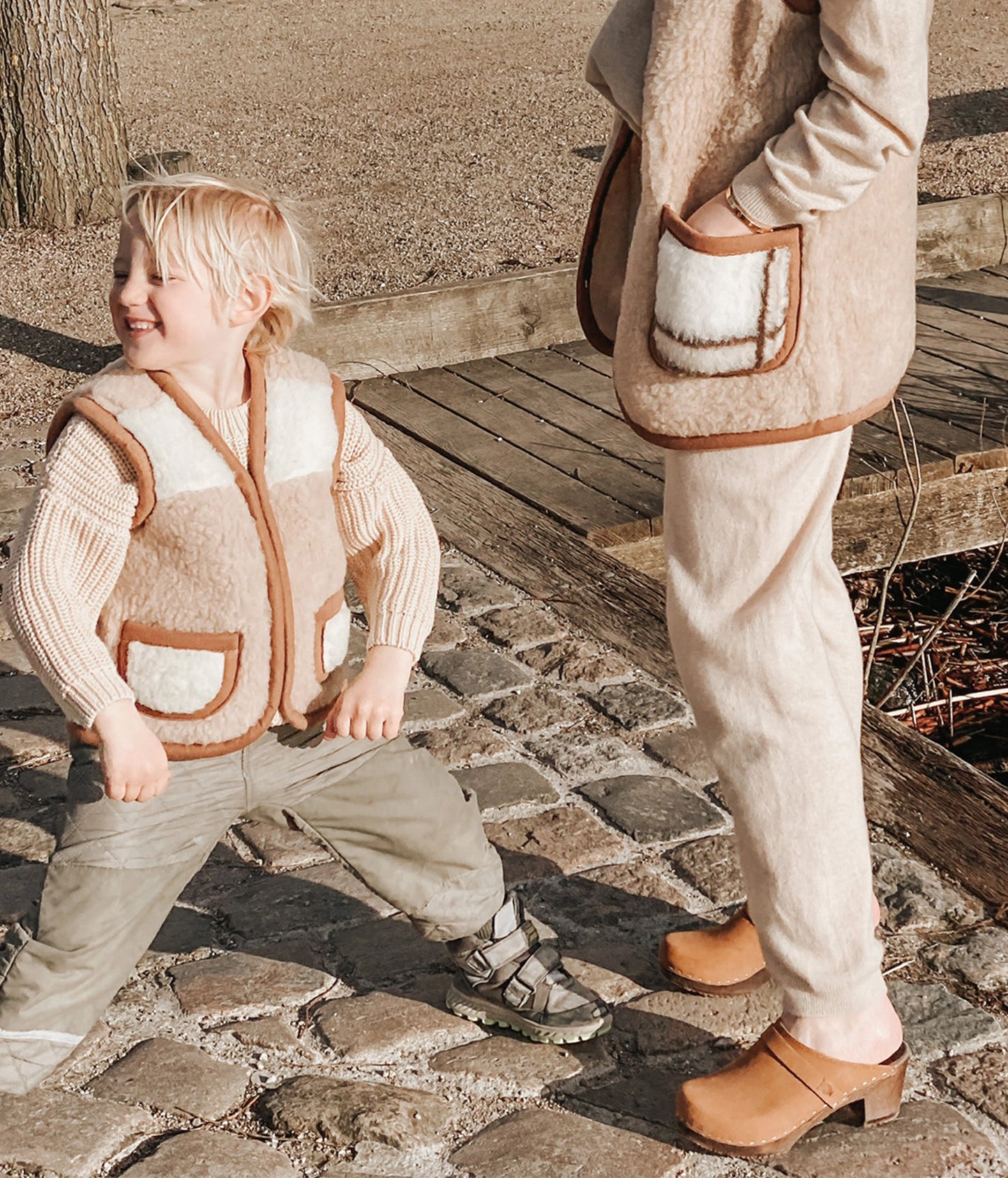 Two children in matching beige outfits standing on a stone path. Classic high heeled clog mule in Dexter tan nubuck leather stapled on a dark wooden base with a leather strap