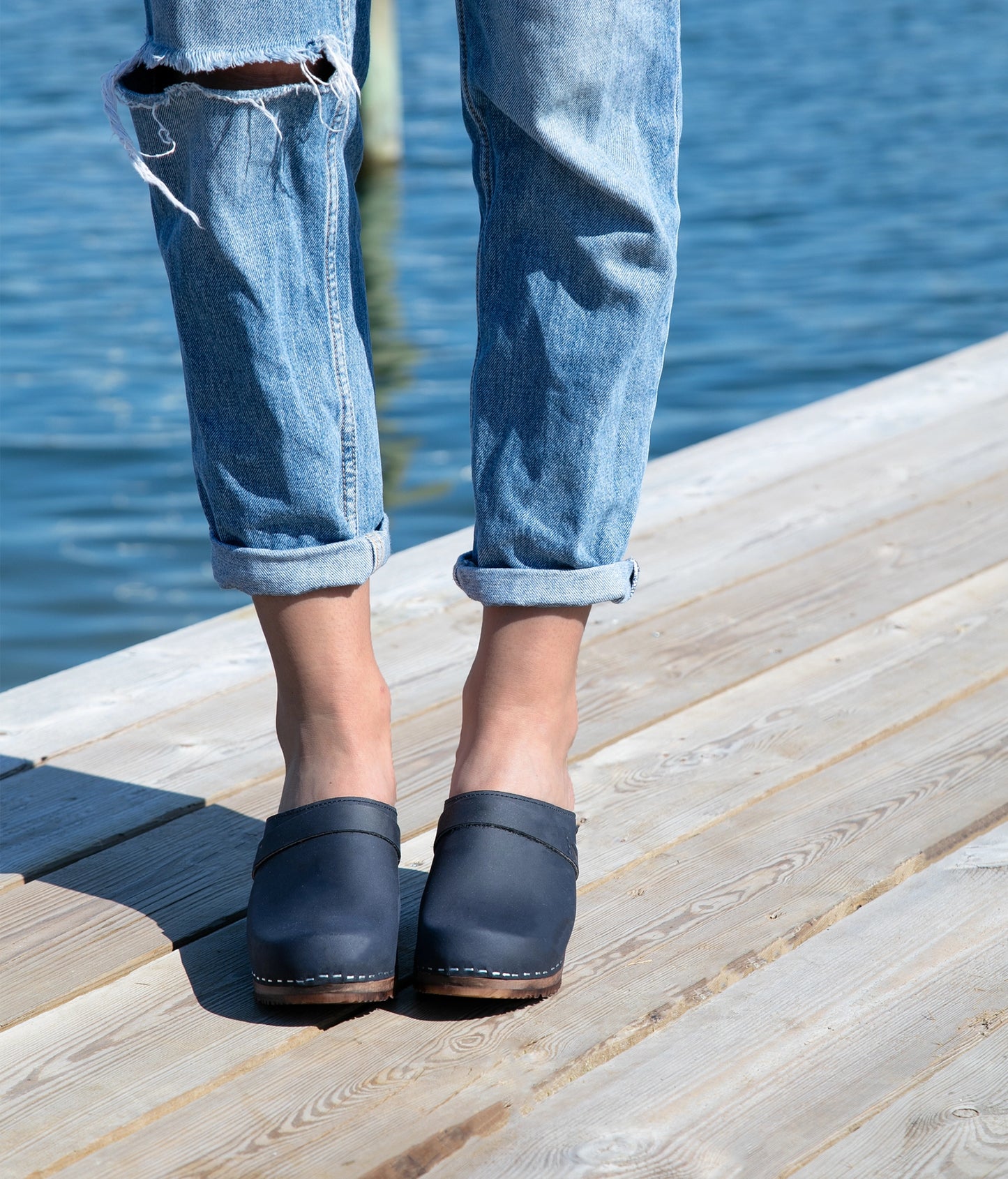 Person wearing blue jeans and navy clogs standing on a wooden dock by water. classic high heeled clog mule in navy blue nubuck leather stapled on a dark wooden base with a leather strap