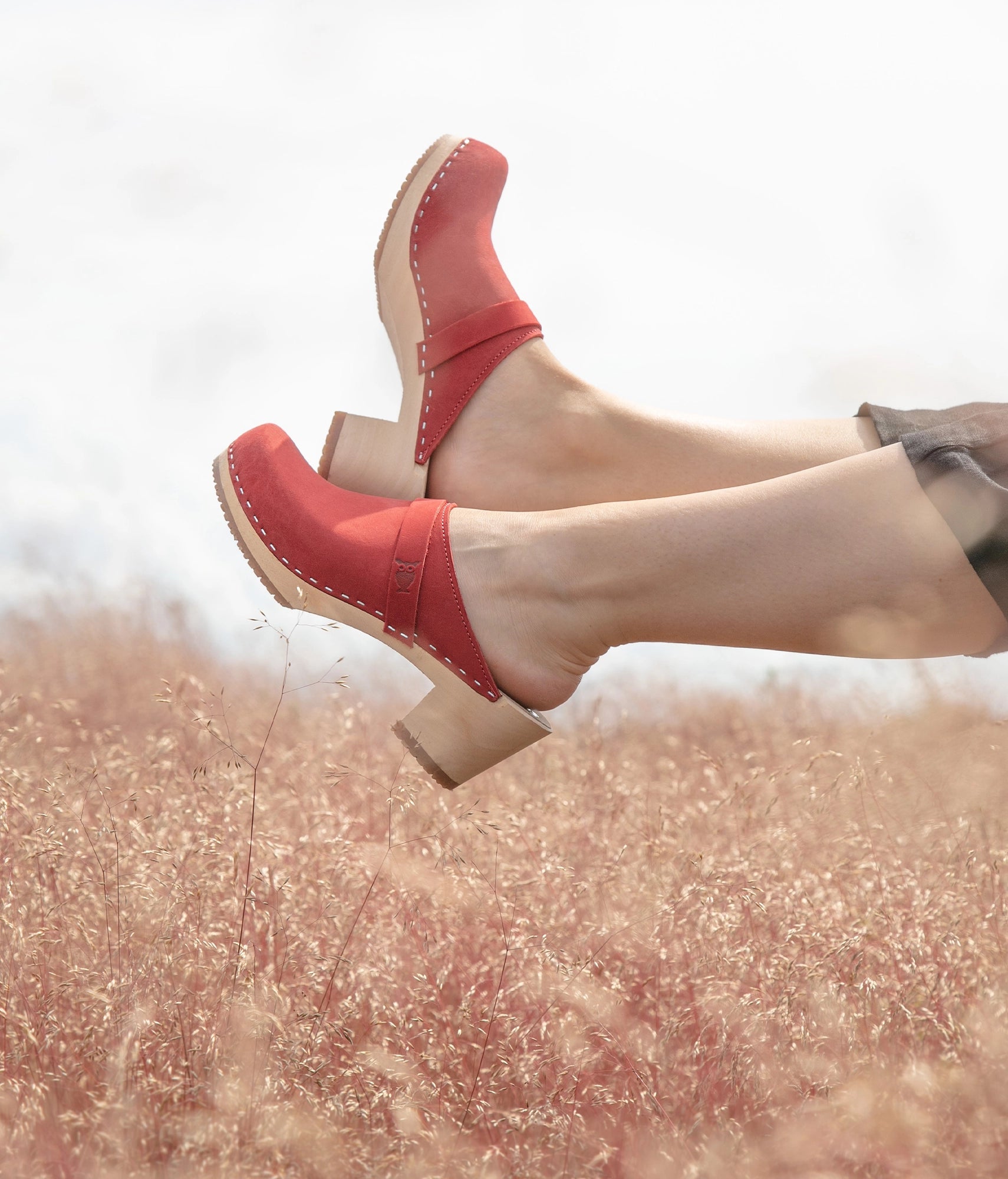 Red clogs on feet with a blurred natural background. classic high heeled clog mule in red nubuck leather stapled on a light wooden base with a leather strap